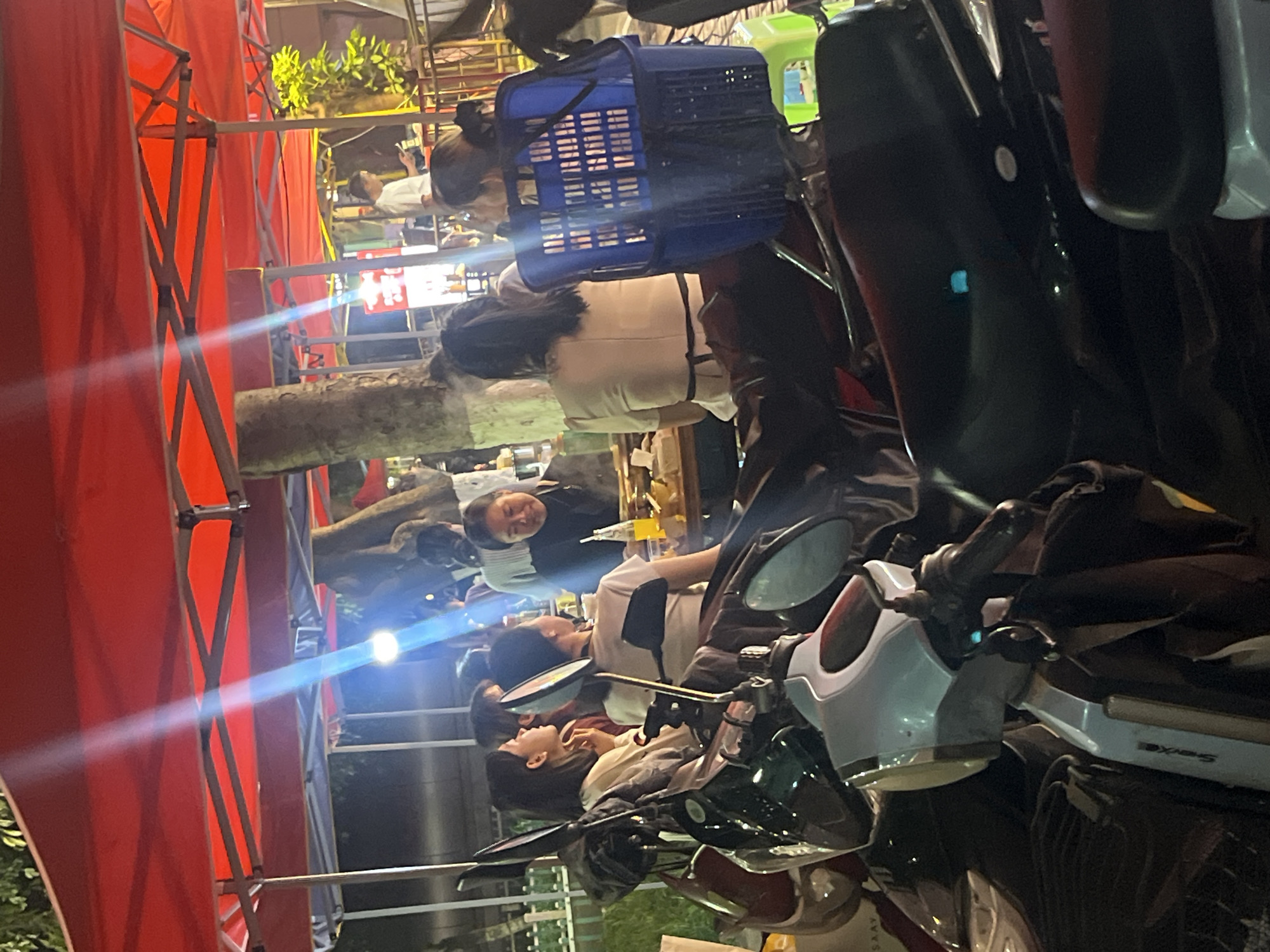 This is a nighttime, medium-distance, slightly low-angle shot of an outdoor dining area in the Yuzhong District of China, possibly a street food market or restaurant. Several individuals are seated at tables under a red canopy. In the foreground, multiple motorcycles are parked, partially obscuring the view of the diners. One person is looking down at their phone, while another is engaged in conversation. A blue plastic basket sits on the back of one of the motorcycles. The lighting is dim, with strong beams of light cutting through the scene from above, possibly from streetlights or overhead lamps. The environment appears to be urban, with trees and other structures visible in the background.