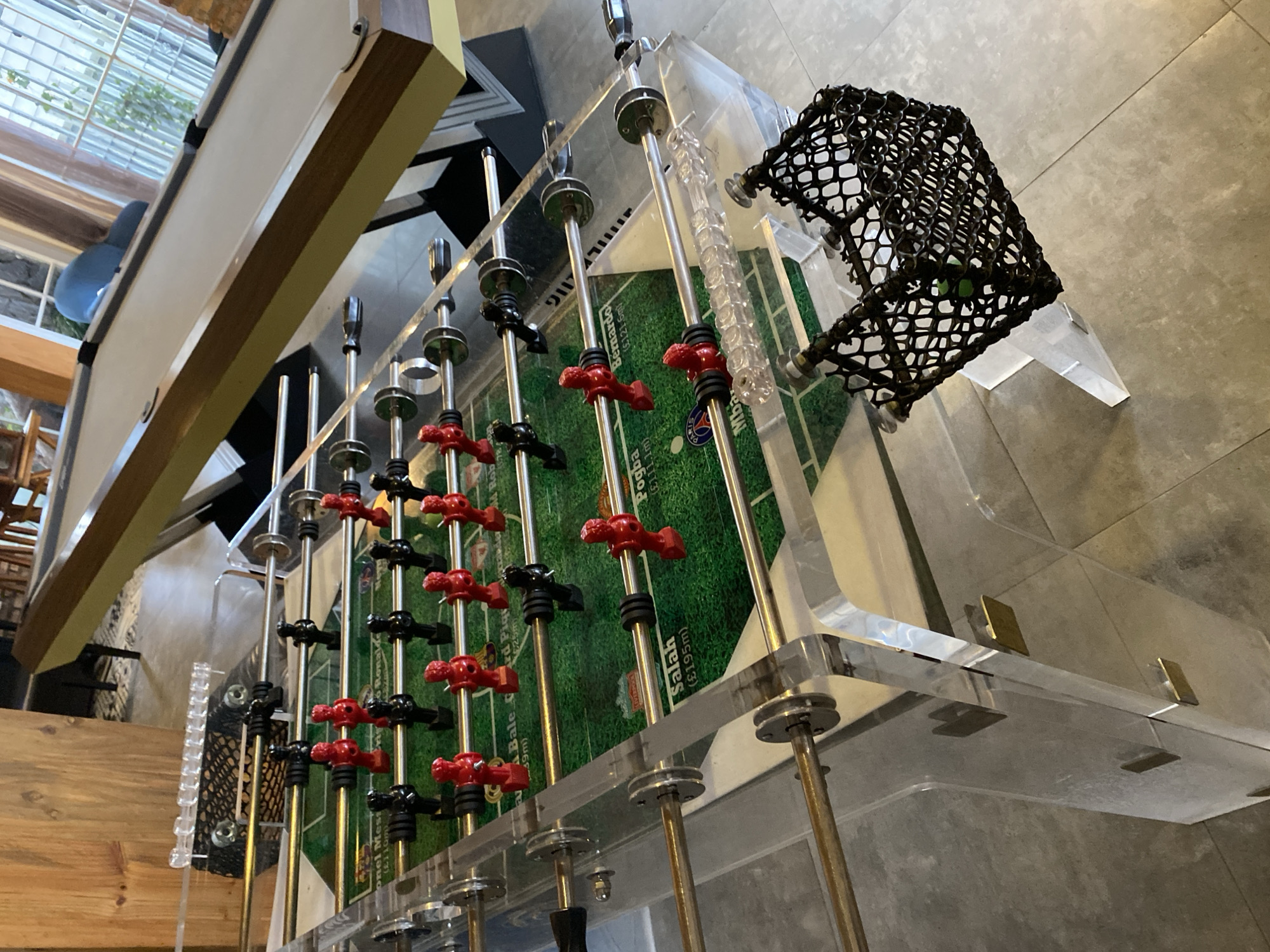This is a close-up, eye-level, medium shot of a modern, transparent acrylic foosball table. The table has green turf with white markings and images of soccer players. The players are depicted in red and black uniforms. Some of the player figures have names and monetary values printed on the turf beneath them, such as "Bale (£39m)" and "Pogba (£111m)." The table is supported by clear acrylic legs, and metal rods extend across it, holding the player figures. Two black mesh nets are positioned at either end of the table, designed to catch the foosball. In the background, a portion of a pool table with a white surface and a wooden frame is visible. The environment appears to be indoors, possibly a recreational area, with wooden paneling and windows with blinds in the far background. The lighting is natural, suggesting daytime. The scene is devoid of any people or animals. It is not possible to determine the location in Da Lat, Vietnam based on the provided image.
