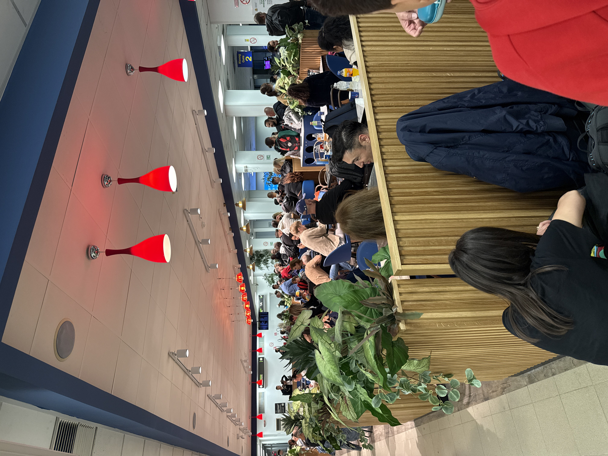 The image depicts a busy indoor public space, likely an airport terminal in Sofia, Bulgaria, during an unspecified time of day. The scene is filled with numerous people, primarily adults, seated at tables and benches, engaged in waiting, conversing, or using electronic devices.
Architecturally, the space features a long, light-colored wall along one side, accented by several distinctive red, cone-shaped light fixtures that cast a warm glow. Above this wall is a blue structural element. A prominent feature on the right side of the image is a tall, vertical wooden slatted wall or partition. Throughout the area, various potted green plants are strategically placed, adding a natural element to the otherwise artificial environment.
In the mid-ground, multiple rows of tables and chairs are occupied by travelers. Many individuals are seen looking down at phones or tablets, while others appear to be interacting with companions. Personal belongings, such as jackets and bags, are visible near the seating. In the background, a blue sign with white text clearly displays "IZHOD Gate 2," indicating "Exit Gate 2" in Bulgarian, confirming the airport context. Another information screen is also visible. The floor is tiled in a light, neutral color. The overall impression is a bustling, functional waiting area, typical of an airport gate.