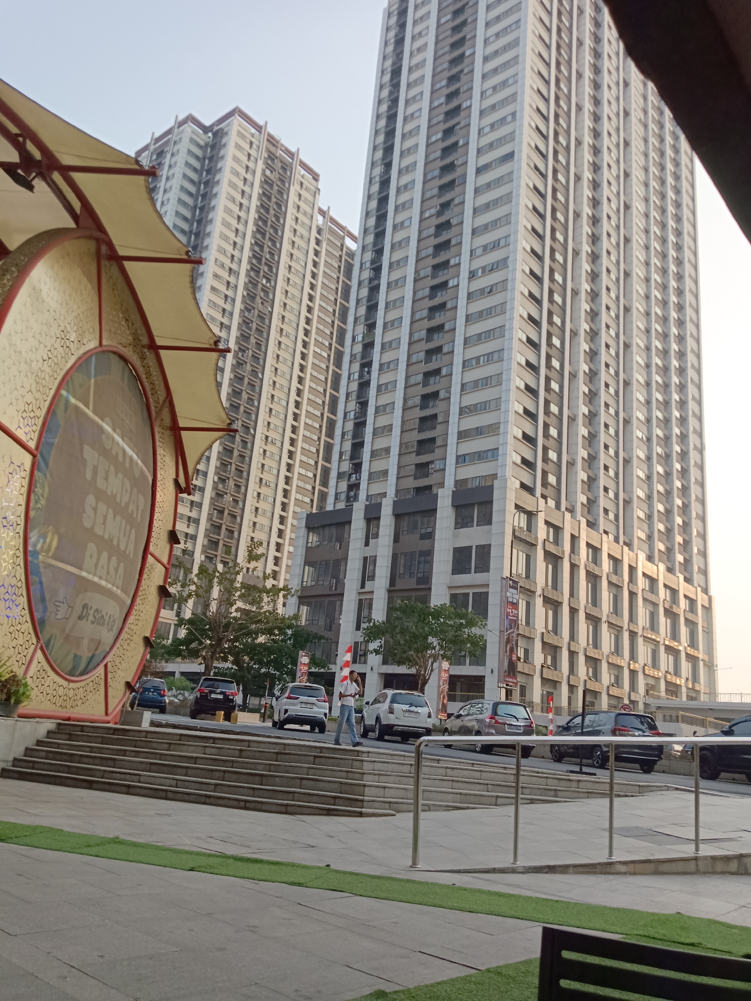 The image depicts a modern urban scene in Jayamukti, Indonesia, dominated by towering residential or office buildings. In the foreground, a large, circular, decorative structure with a beige canopy and intricate golden detailing is visible on the left. The structure has text within its circular opening that reads "SATU TEMPAT SEMUA RASA" and "Di Sini Ra". To the right of this structure, a broad set of stairs leads up to a street level where several cars are parked. Among the parked vehicles are a white SUV, a dark sedan, and a darker SUV. A man in a light-colored shirt and blue jeans is walking up the stairs. Behind the stairs, more cars are visible on a slightly elevated road, including a dark SUV and a black sedan. Beyond the cars and stairs, the imposing facades of multiple high-rise buildings rise into the sky, characterized by numerous windows and a light-colored exterior. Green grass patches are visible in the lower part of the frame. The sky is a hazy, pale color, suggesting it is either early morning or late afternoon.