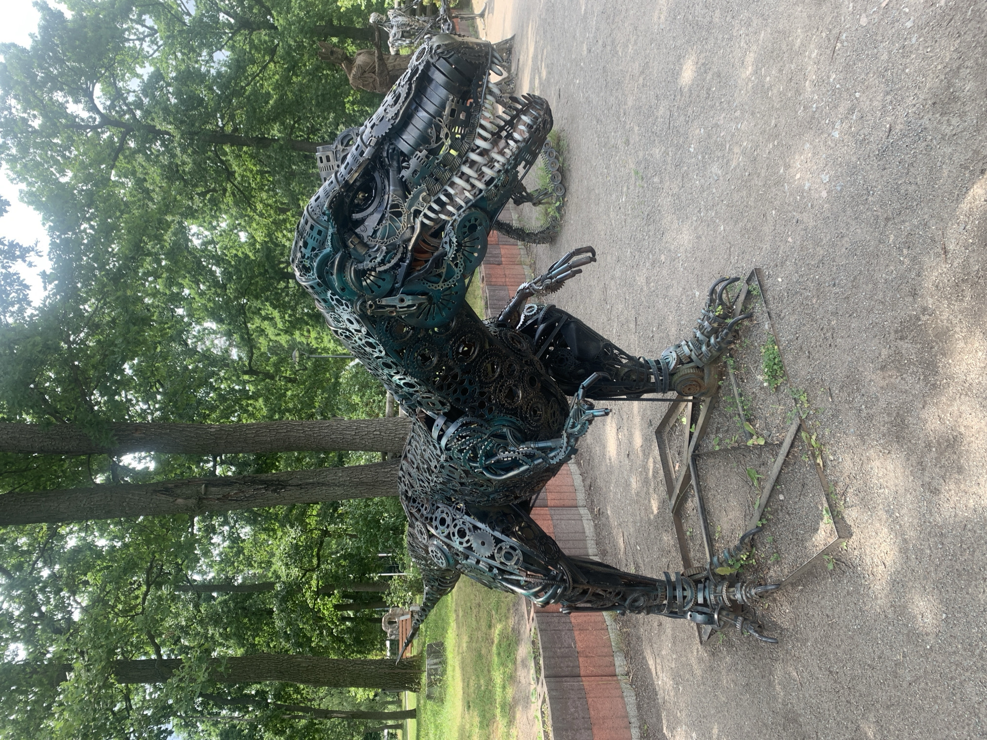 This image depicts a large, metallic sculpture of a Tyrannosaurus Rex, likely made from recycled metal parts such as gears and chains. The sculpture is dark green and black, with exposed metal components giving it a mechanical, almost skeletal appearance. It stands on a flat, square metal base surrounded by gravel. The T-Rex sculpture is situated outdoors, with a backdrop of lush green trees and a partly overcast sky. To the right of the T-Rex, other metallic sculptures are visible, including a humanoid figure and another dinosaur-like creature. In the background, there is a low brick wall and some grassy areas. The overall setting appears to be a park or an outdoor exhibition space. This scene is located in the city of Bucha, Ukraine.