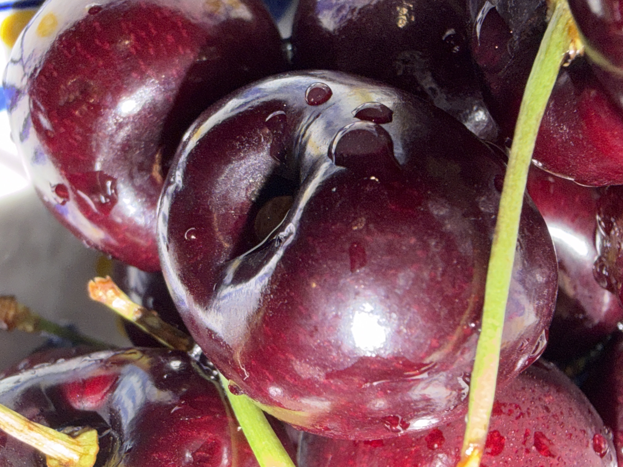 A close-up, high-angle, macro photograph shows a pile of ripe, dark red cherries. The cherries are wet, with visible water droplets on their surfaces, indicating they are fresh and possibly recently washed. Some of the cherries have stems attached, which are a vibrant green color. The lighting creates strong highlights and reflections on the glossy skin of the cherries, emphasizing their plumpness and deep, rich color. The background is dark and out of focus, serving to draw attention to the cherries. The image is a still life, focusing purely on the visual appeal of the fruit.
