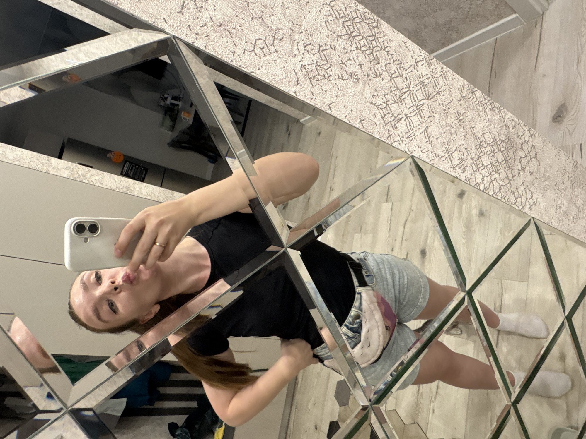 A woman is taking a selfie in a mirror with a geometric design, possibly composed of multiple smaller mirrors, in Izhevsk, Russia. She's holding a white smartphone, puckering her lips, and wearing a black shirt, jean shorts, and white socks. The reflection in the mirror shows parts of a room with a patterned wall and floor.