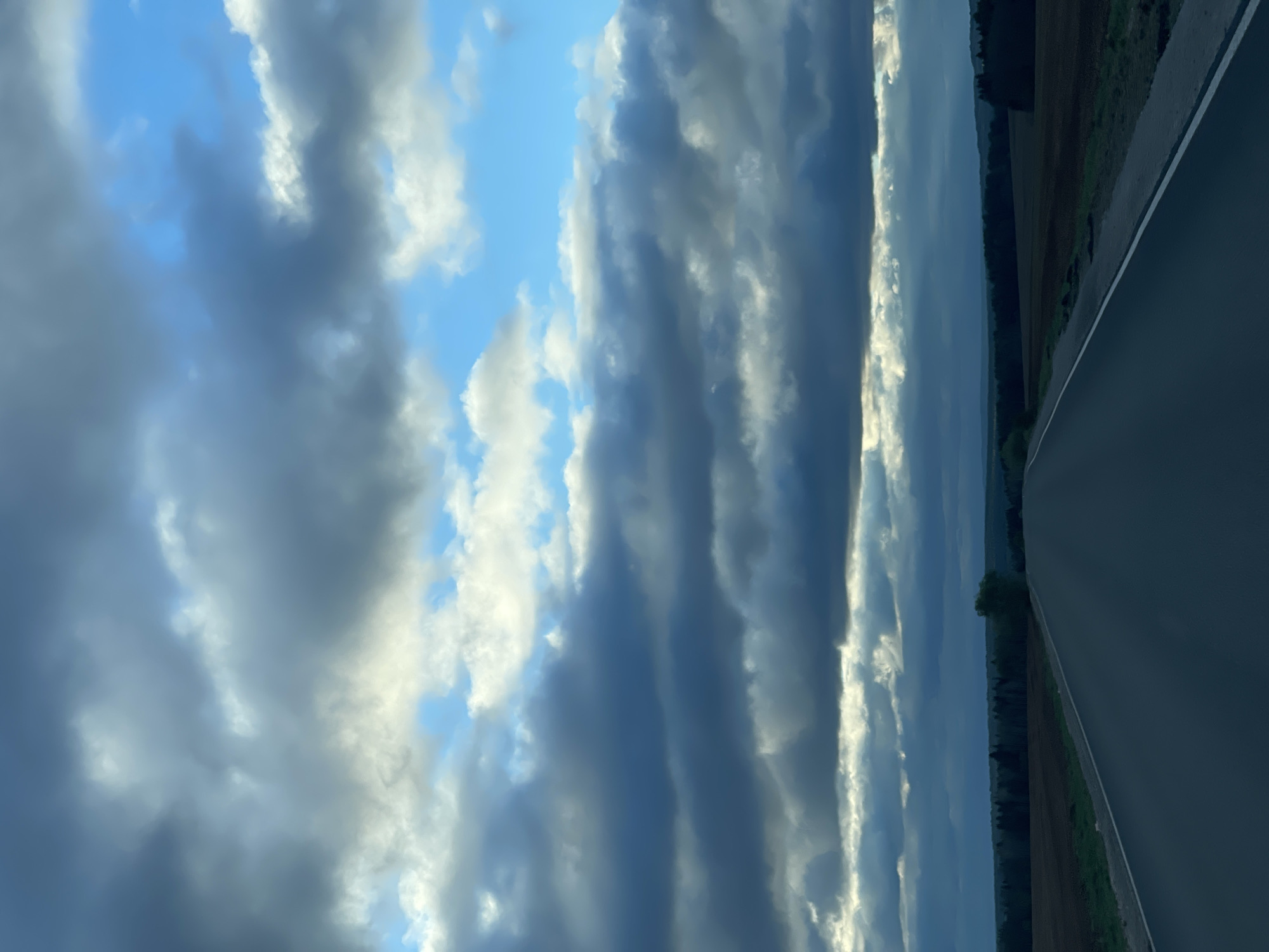 The image shows a scene from what appears to be a road near Перевозное, Russia, though this cannot be definitively confirmed. The primary view is of a sky filled with layered, striated clouds, ranging from light grey to dark grey, with patches of blue sky visible. A road runs along the right side of the frame. Along the road is a strip of land with trees and what appears to be fields. The light indicates it may be late afternoon or early evening. There are no people visible in the image. The focus is on the cloudscape and the landscape alongside the road.