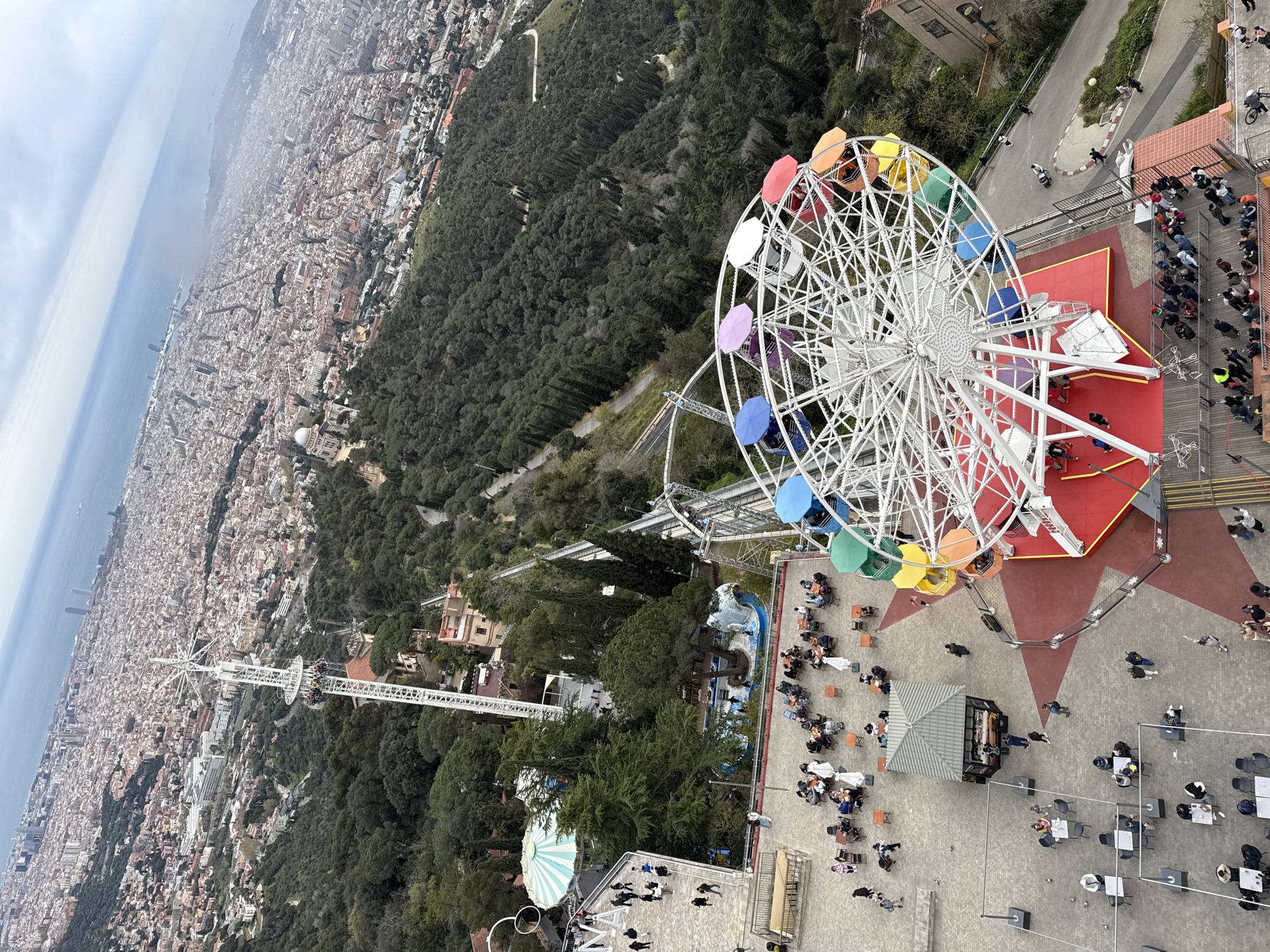 The image shows an aerial view of Tibidabo Amusement Park in Barcelona, Spain. A colorful Ferris wheel is a prominent feature. Below, people are seated at tables and queuing in a roped off area. A roller coaster and other amusement park rides are visible, surrounded by dense trees. In the distance, the city of Barcelona stretches to the sea under a cloudy sky.