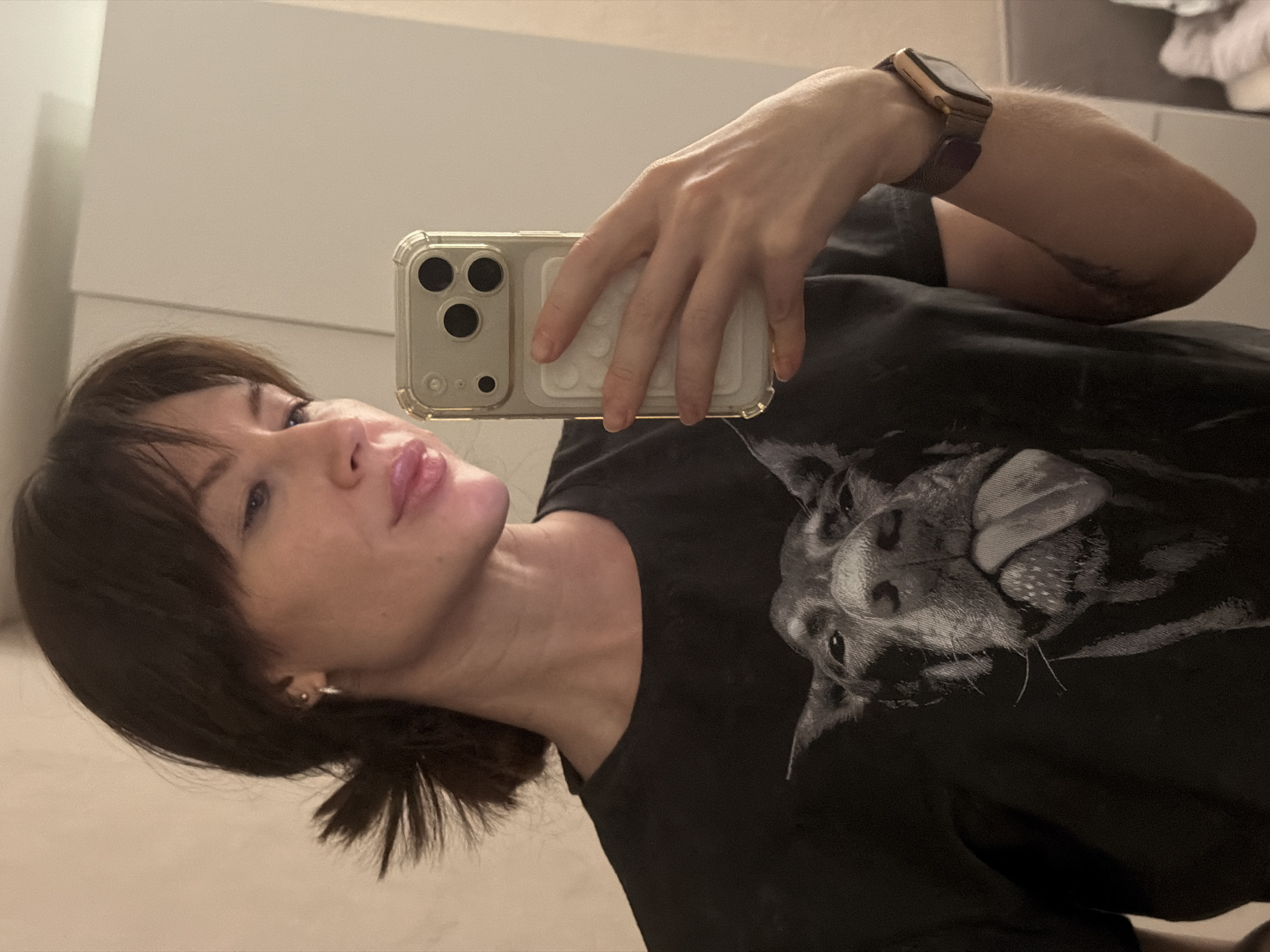 A woman is taking a selfie in what appears to be an indoor setting. She is holding a smartphone in a clear case. She is wearing a black t-shirt with a dog printed on it and a watch. The background is out of focus, but what appears to be white furniture can be seen. The location is in Perm, Russia.