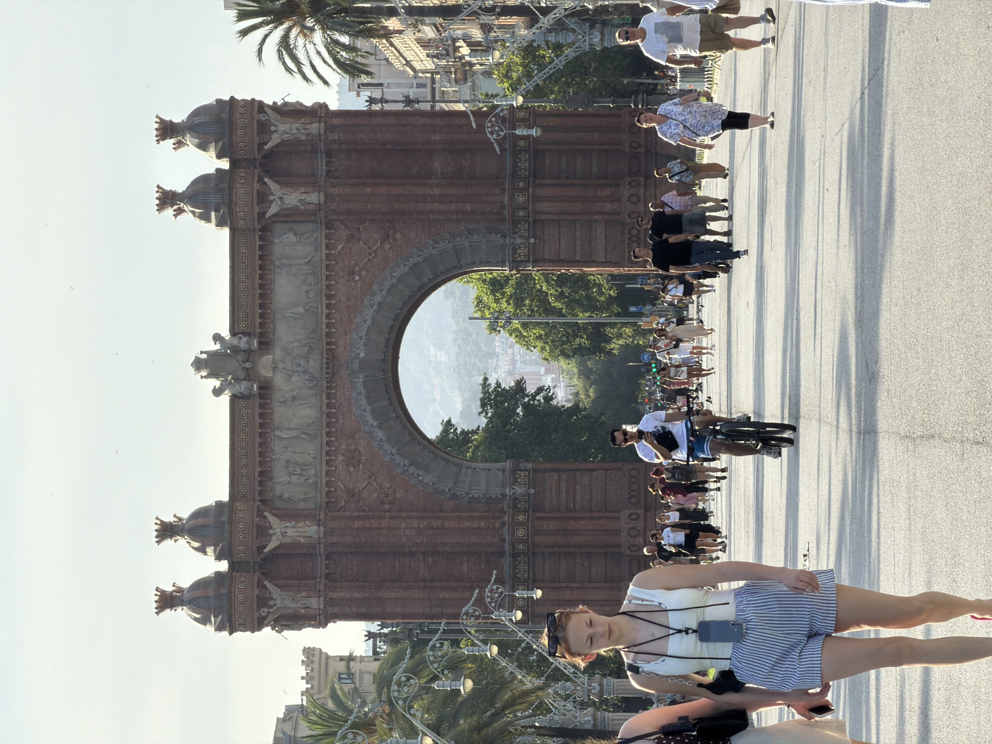 The image captures a view of the Arc de Triomf in Barcelona, Spain, under a bright, hazy sky. The monumental brick arch dominates the background, featuring ornate carvings and topped with decorative domes crowned with smaller spires. In front of the arch, a wide, paved walkway is bustling with people.
In the foreground, a young woman with blonde hair pulled back and sunglasses perched on her head walks towards the camera. She is wearing a white tank top and blue and white striped shorts. She has a lanyard with a phone case around her neck. To her left, a sliver of another person is visible.
Further down the walkway, a man on a bicycle rides away from the camera, wearing a white t-shirt and blue shorts. He is looking at his phone. To his right, a group of people are walking, including a woman in a white and blue floral print top and black leggings, and a man in a white t-shirt and beige shorts.
The walkway is lined with palm trees and decorative lampposts with multiple lights. The overall scene depicts a lively public space with tourists and locals enjoying a sunny day near a significant city landmark.