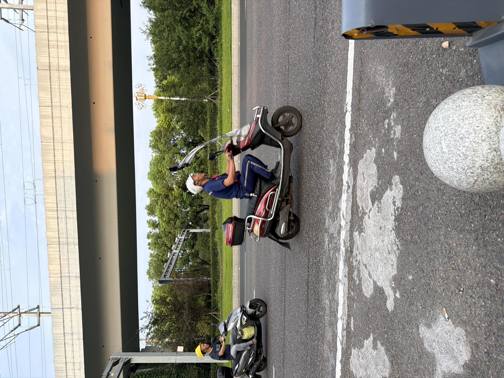 In Jiangjin, China, two individuals are riding scooters on a paved road beneath an elevated overpass. The person on the left, wearing a yellow hard hat and grey clothing, rides a silver scooter with a green bag attached. The person on the right, wearing a white helmet and blue work uniform, rides a red and black scooter with a black storage box on the back. The environment includes trees and greenery lining the road, overhead power lines, and a street lamp in the background. The road surface shows a white lane marking and some water stains. A discarded cigarette butt is visible on the road in the foreground, near a spherical decorative element and a traffic safety bollard.