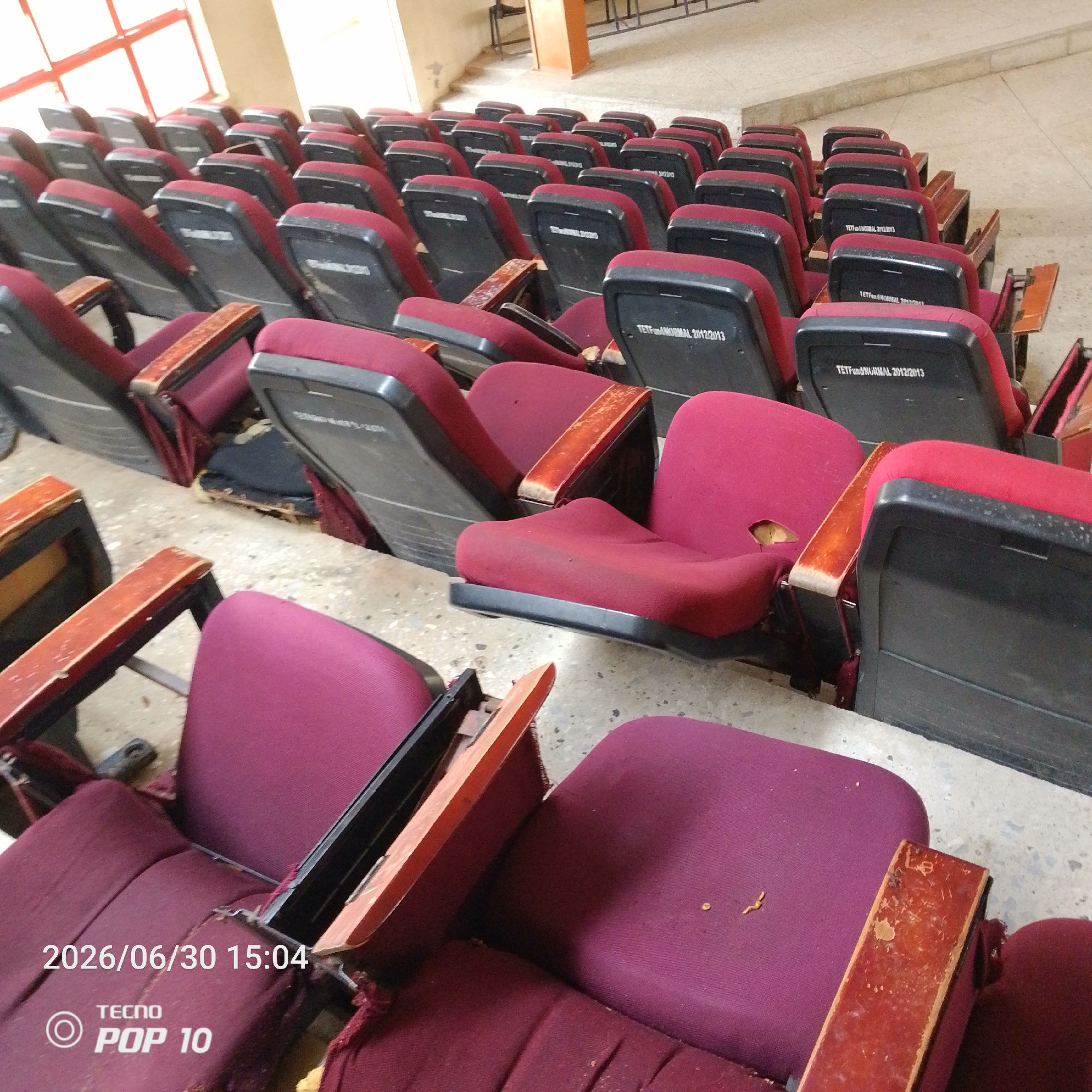 The image shows rows of empty, maroon upholstered seats with black plastic backs in what appears to be an auditorium or lecture hall. Some of the seats show signs of wear and tear, with worn armrests and ripped upholstery. The floor is concrete, and a window with red-framed panes is visible in the upper left corner, letting in bright light. The lighting suggests it is daytime.
Several seats have text visible on the back, including "TETFund/NORMAL 2012/2013" and "TETFNORMAL 2012/2013". In the bottom left corner, a timestamp and brand logo are visible: "2026/06/30 15:04" and "TECNO POP 10". There are no people visible in the image. The scene is static and quiet, suggesting the room is not in use at the moment the photo was taken. The overall impression is of a functional, but somewhat aged, institutional space.