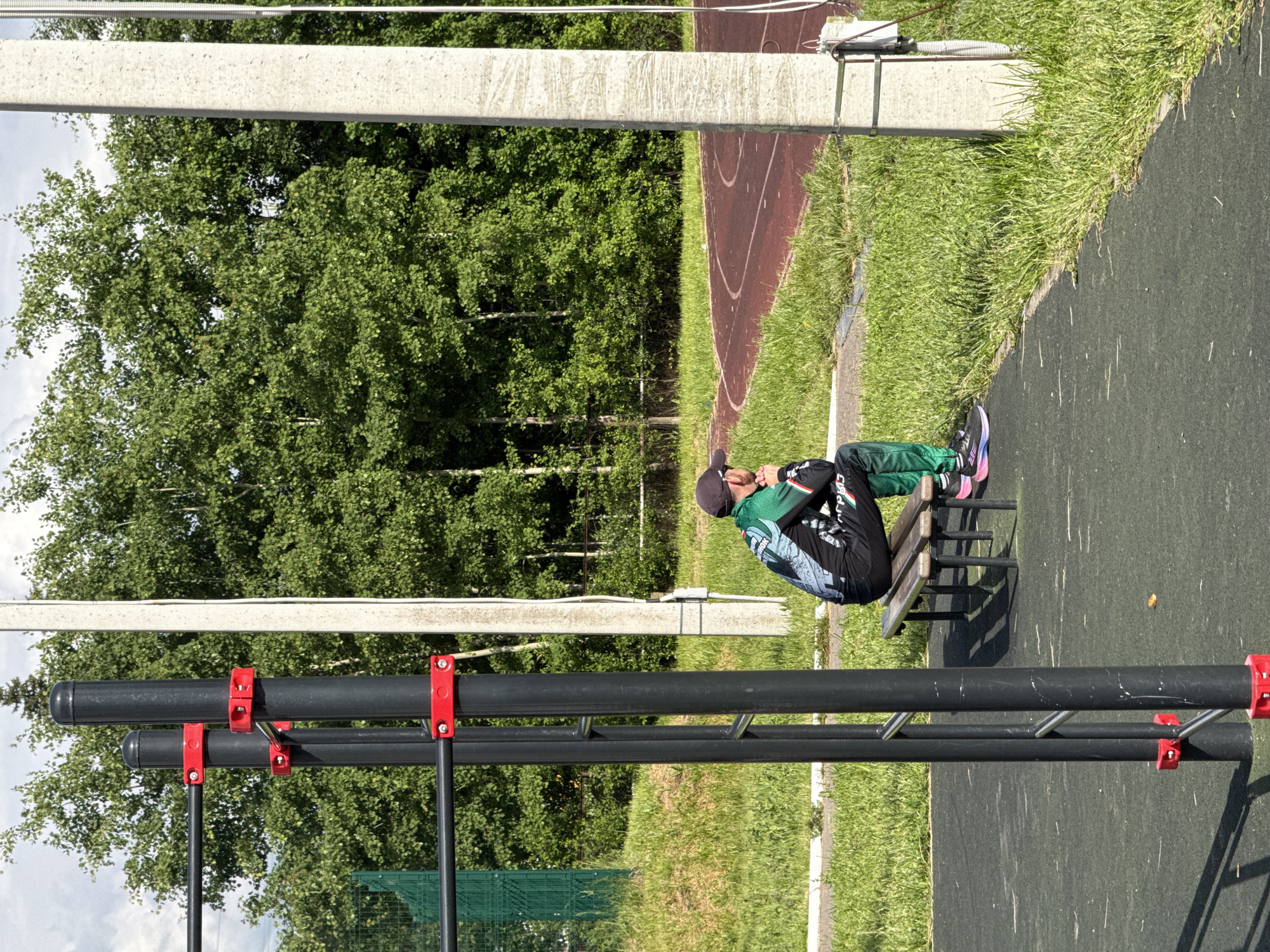 This image depicts an outdoor fitness area or sports ground, likely located in Прикамский, Russia. The photograph is rotated 90 degrees clockwise, presenting the scene vertically.
In the midground, a single person, appearing male, is seated on a light-colored bench. They are dressed in athletic wear, including a dark cap, a black and green long-sleeved top with distinctive white patterns, and dark athletic pants. The person is shown leaning forward slightly, with hands clasped around their knees, in what appears to be a resting or contemplative posture. Their athletic shoes have noticeable bright pink soles.
In the immediate foreground and to the left of the person, there are black metal outdoor exercise structures. These include parallel bars and a multi-height pull-up or ladder-style station, all featuring striking red clamps and connectors. The ground directly beneath the exercise equipment and the person is a dark grey or black rubberized surface, typical of outdoor gyms.
The background is dominated by lush green deciduous trees, possibly birch, forming a dense natural screen. Beyond a strip of vibrant green grass, a section of a red running track with white markings is visible, suggesting the area is part of a larger sports complex. The sky above is bright with some light clouds, indicating a clear, sunny day.
Two light-colored concrete pillars or beams are present: one on the far left edge and another running horizontally across the upper part of the image, potentially part of an overhead support structure or fence.
Visible text on the person's athletic top includes the Cyrillic word "АГРО" on the right sleeve and what appears to be "СПОРТ" (SPORT) on the left shoulder/back area of the garment.