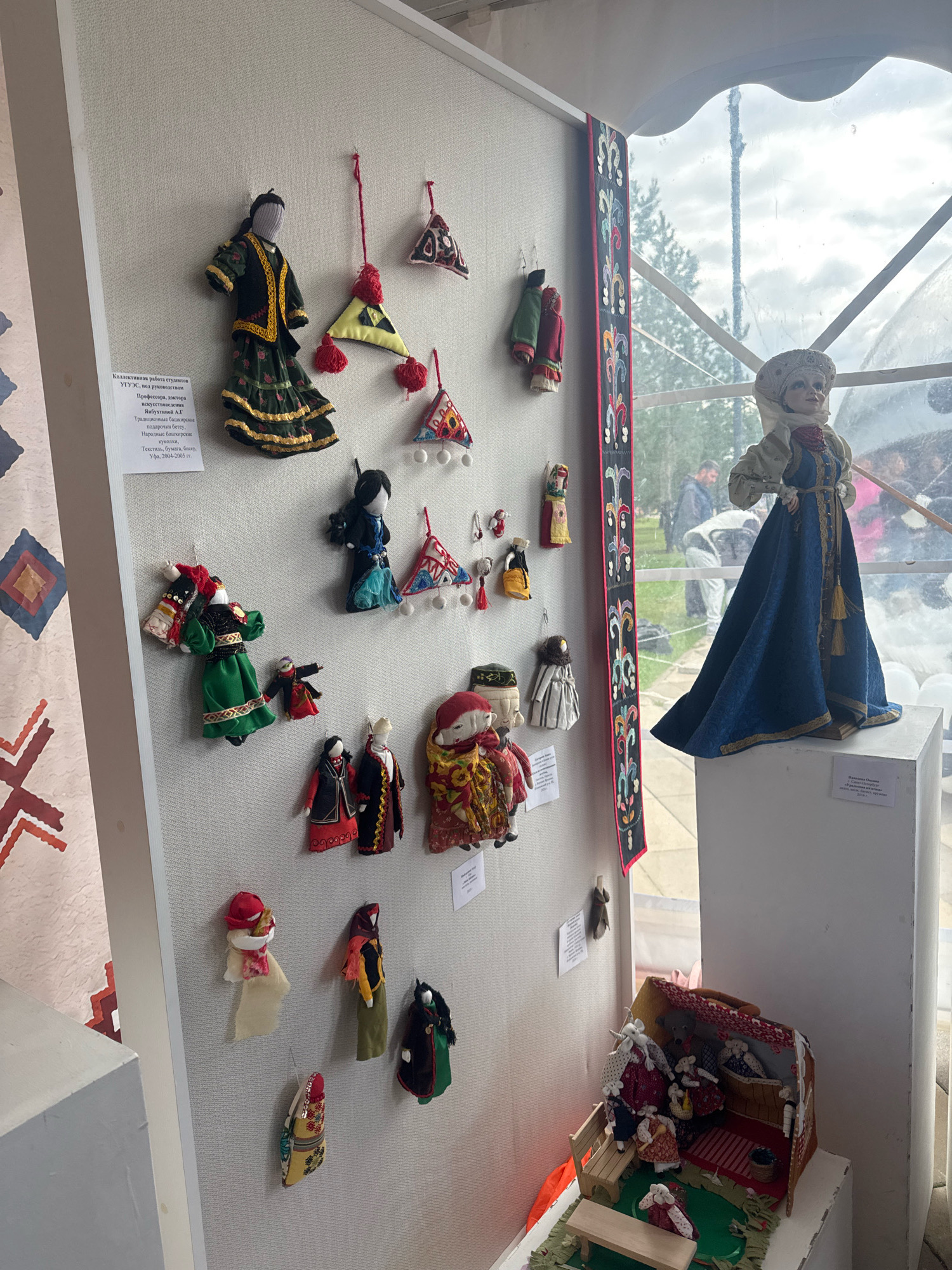 The image depicts an exhibition of traditional Bashkir dolls in Ufa, Russia. The display features numerous handmade dolls of various sizes and designs, showcasing intricate traditional clothing and accessories. Some dolls are hung on a textured white wall, while others are arranged in a miniature diorama of a house, complete with furniture and tiny inhabitants. A large doll in a blue dress stands prominently on a pedestal to the right of the display. In the background, through a large window, glimpses of trees and people can be seen, suggesting the exhibition is held in a public space with natural light. The overall scene is one of cultural exhibition and artistic craftsmanship.