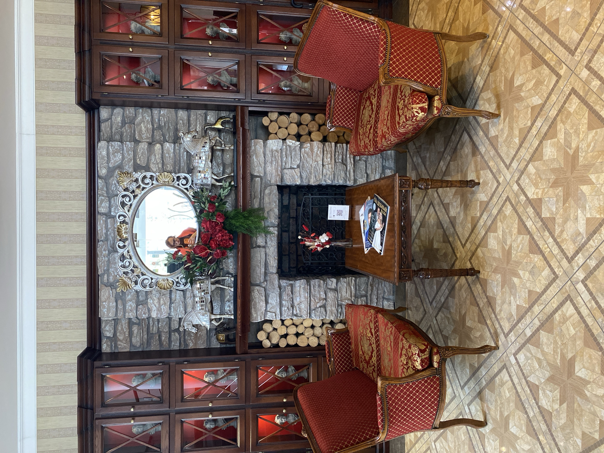The image depicts an interior space in Yekaterinburg, Russia, featuring a decorative fireplace with a stone surround. The fireplace is flanked by built-in wooden cabinets with glass doors, displaying sculptural objects. Above the fireplace is an ornate oval mirror, reflecting a person in an orange shirt. A floral arrangement adorns the fireplace mantel. Two red upholstered chairs with ornate wooden frames face each other, with a small wooden table positioned between them. Magazines and decorative items rest on the table. The floor is covered with geometric patterned tiles.