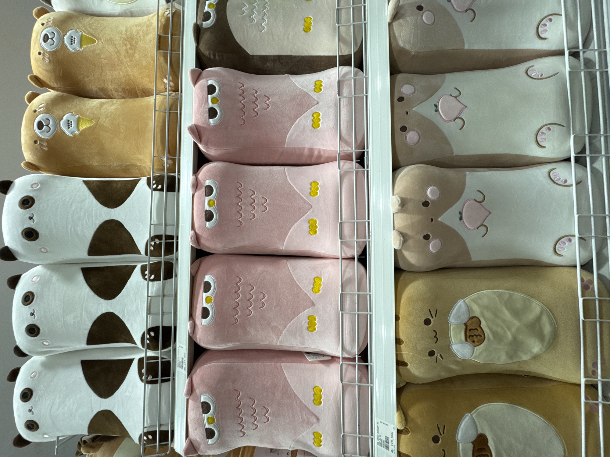 The image shows a retail display of plush pillows in Kuta Selatan, Indonesia. The pillows are arranged vertically on shelves behind a white metal grid. The pillows are of various designs, including a bear with an ice cream cone, a panda, an owl, and a cat with a peach motif. The environment appears to be indoors, likely within a store. There are no people visible in the image. The lighting is diffused and even.