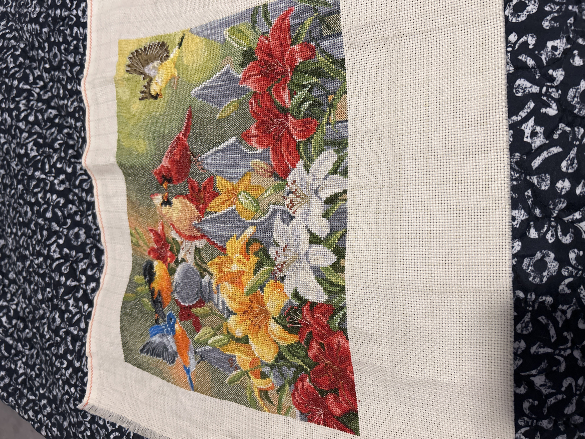 The image shows a partially completed cross-stitch of a floral and bird scene. The stitching depicts several birds, including what appear to be cardinals, a blue and orange bird, and a yellow bird in flight. These birds are positioned among various lilies, with red, yellow, and white blooms. The background features grey mountainous shapes and a green, possibly grassy, area. The cross-stitch is on a light-colored fabric with frayed edges, and it is laid on top of a dark fabric with a white floral pattern. There are no people or specific city landmarks visible in the image.
