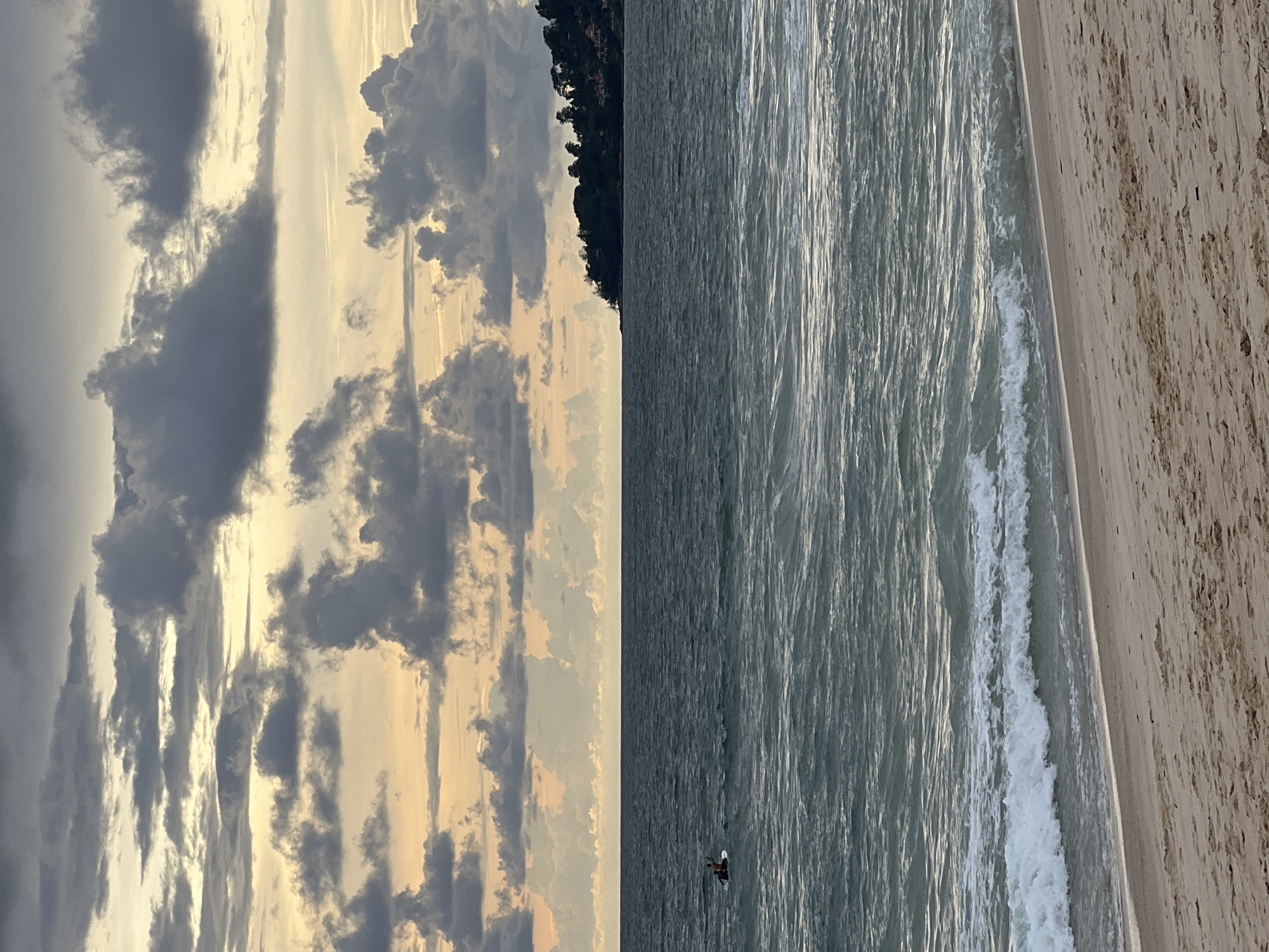 The image depicts a serene beach scene at dusk. The sky is a dramatic canvas of dark, textured clouds with streaks of warm, golden light breaking through, creating an atmospheric and slightly moody ambiance. The ocean is a deep, greenish-blue, with moderate waves lapping against the sandy shore. In the distance, a small boat with two occupants is visible on the water, a lone surfer on a paddleboard in the middle distance, and a lush, tree-covered headland juts out into the sea on the right, dotted with scattered buildings. The foreground is a sandy beach, appearing damp and with subtle ripples from the receding tide. The overall impression is one of tranquil natural beauty, possibly at the end of a day.