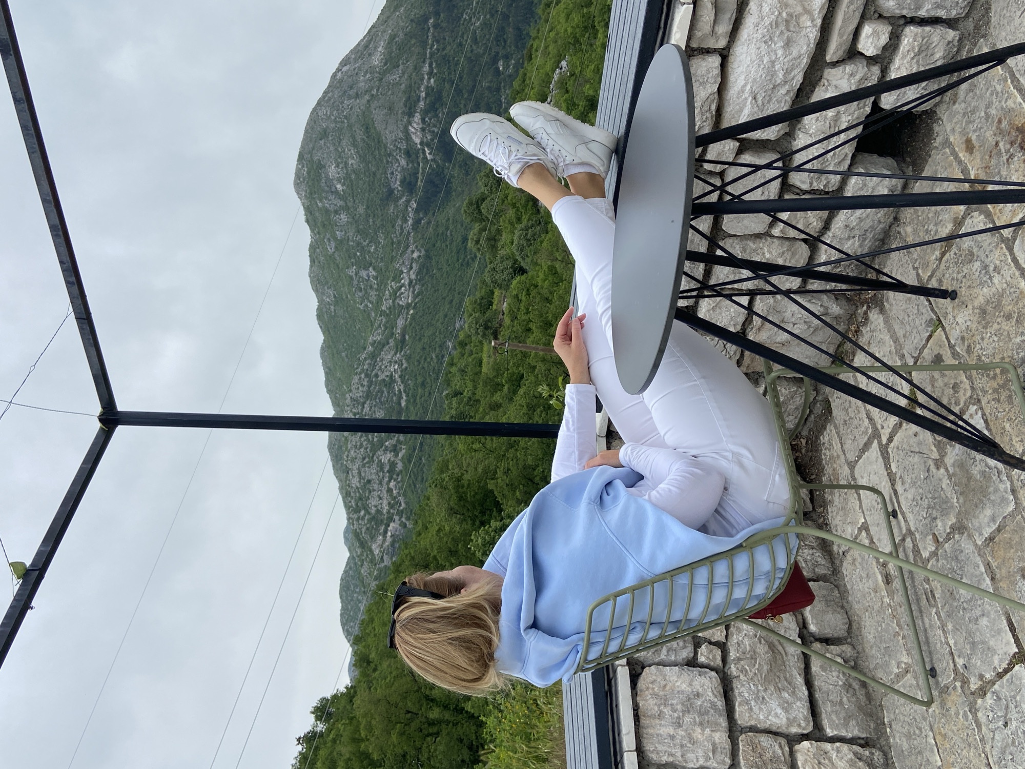 A woman in white pants and white sneakers, wearing a light blue hoodie, is sitting backwards in a green chair with her feet propped on a black metal table. She is looking out at a mountainous landscape. The scene is set on a stone patio with a metal pergola above. The location is in Lipa Dobrska, Montenegro.