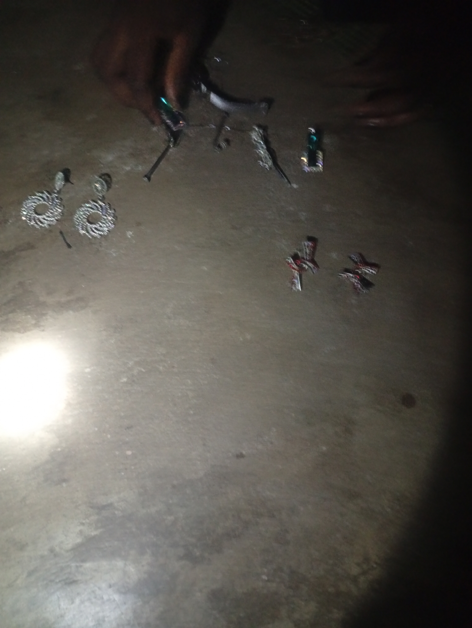 The image depicts a dark, gritty outdoor scene, likely a street or courtyard, in Jalingo, Nigeria. The ground appears to be dirt or rough concrete, illuminated by a bright light source, possibly a flashlight, in the lower left quadrant. Several objects are scattered on the ground. In the upper center, a person's hands are visible, manipulating small, metallic objects, some of which have a green or iridescent sheen. Among these objects are what appear to be pieces of jewelry or decorative items. To the left of this area, two intricately designed silver-toned hoop earrings are laid out. Further to the right, near the center of the frame, are three small, cross-shaped pendants or charms, with red and dark metallic accents. The overall impression is of personal belongings or trinkets laid out on a rough surface in a dimly lit environment, possibly being sorted or examined.