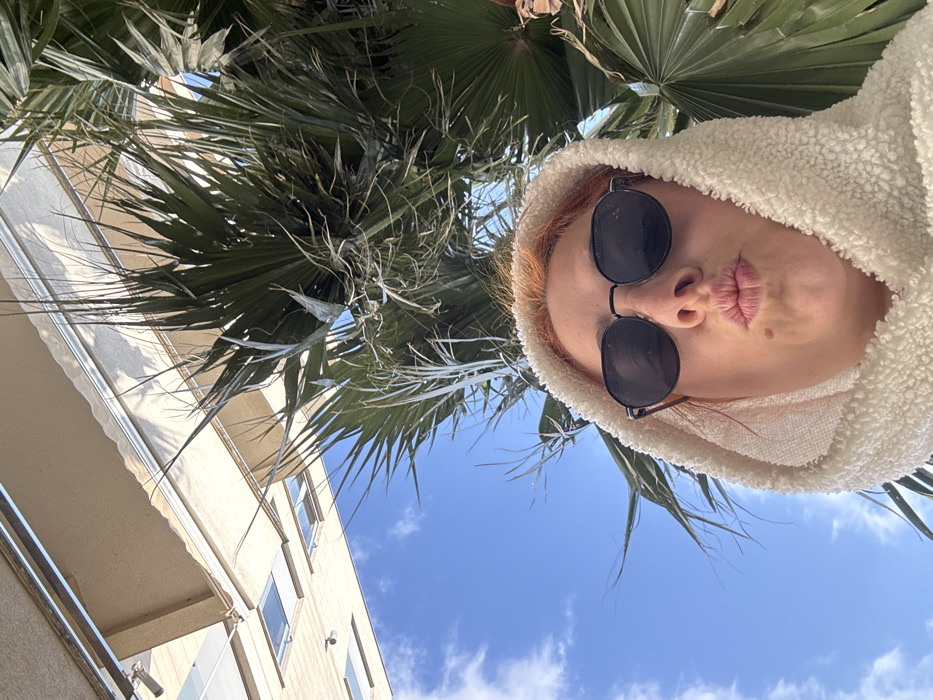 A low-angle selfie image depicts an adult female positioned in the lower-right, wearing dark, circular-framed sunglasses and an off-white, textured hooded garment. Her reddish-brown hair is visible above her forehead, and she has a mole below her right lower lip. Her lips are pursed in a "kissing" gesture, directed towards the camera.
The background features a clear blue sky with scattered white clouds. To the right and behind the subject, dark green fronds of a fan palm tree are visible. In the upper-left quadrant, a light-beige multi-story building with multiple rectangular windows and an extended awning appears inverted, suggesting an upward-facing camera orientation. Small light fixtures are mounted on the building's facade. The scene is illuminated by direct sunlight, indicating an outdoor, daytime setting in Budva, Montenegro.