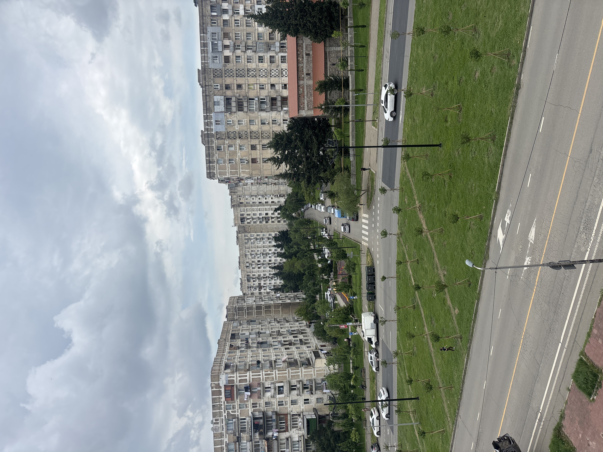 The image depicts a street scene in Tbilisi, Georgia, characterized by Soviet-era apartment buildings. The architecture is marked by repetitive, multi-story concrete structures with numerous windows and balconies. A multi-lane road runs parallel to these buildings, with vehicles moving in both directions. The road is flanked by grassy medians planted with saplings. A pedestrian crossing is visible. The sky is overcast with grey clouds. A street light is on the right side of the road.