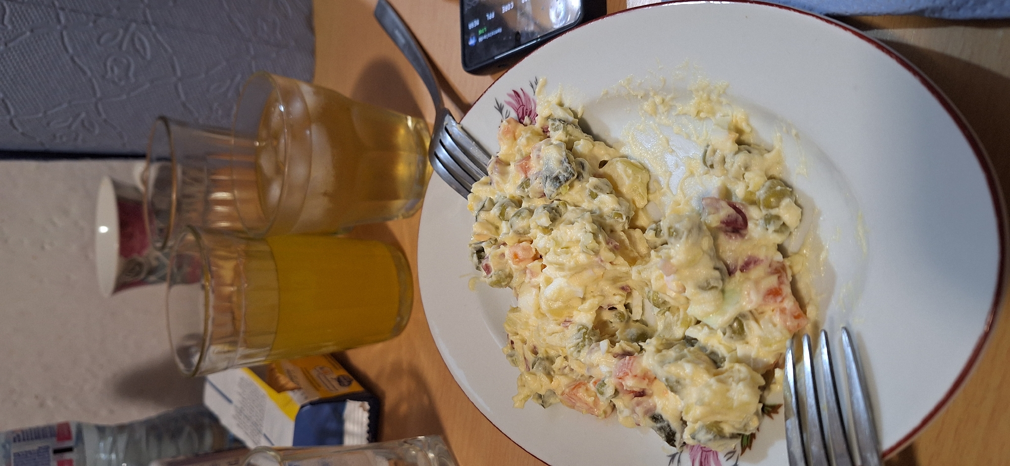 The image displays an indoor scene, likely a dining table, set for a casual meal. The primary subject is a white plate, bordered with a red rim and a floral design, containing a generous serving of a creamy mixed vegetable salad. This salad appears to be a variation of Olivier salad, which is common in the region, featuring diced potatoes, peas, carrots, and other ingredients like pickles or possibly meat, all coated in a mayonnaise-based dressing. A silver fork is visible resting on the plate, with tines submerged in the salad.
To the left of the plate, two clear glass tumblers hold beverages. The bottom glass contains a vibrant orange liquid, while the top glass holds a lighter yellow drink. A dark-colored smartphone lies face up near the top left of the plate, with its screen visible. Text such as "CORE APL MENU" and "ORIGINAL" can be discerned on the screen amidst various icons. Another silver fork is seen near the smartphone, positioned between it and the salad plate.
Further left in the background, a white cup with a faded red and green floral pattern is partially visible. A plastic water bottle and a blue and white snack package are also present on the table, though their labels are mostly obscured. The table surface appears to be a light wood. The background consists of a light-colored wall with a subtle textured pattern.
The scene depicts a paused meal or snack. No people are visible. The lighting suggests an indoor setting, but the specific time of day is not discernible. The overall impression is one of a simple, everyday meal.