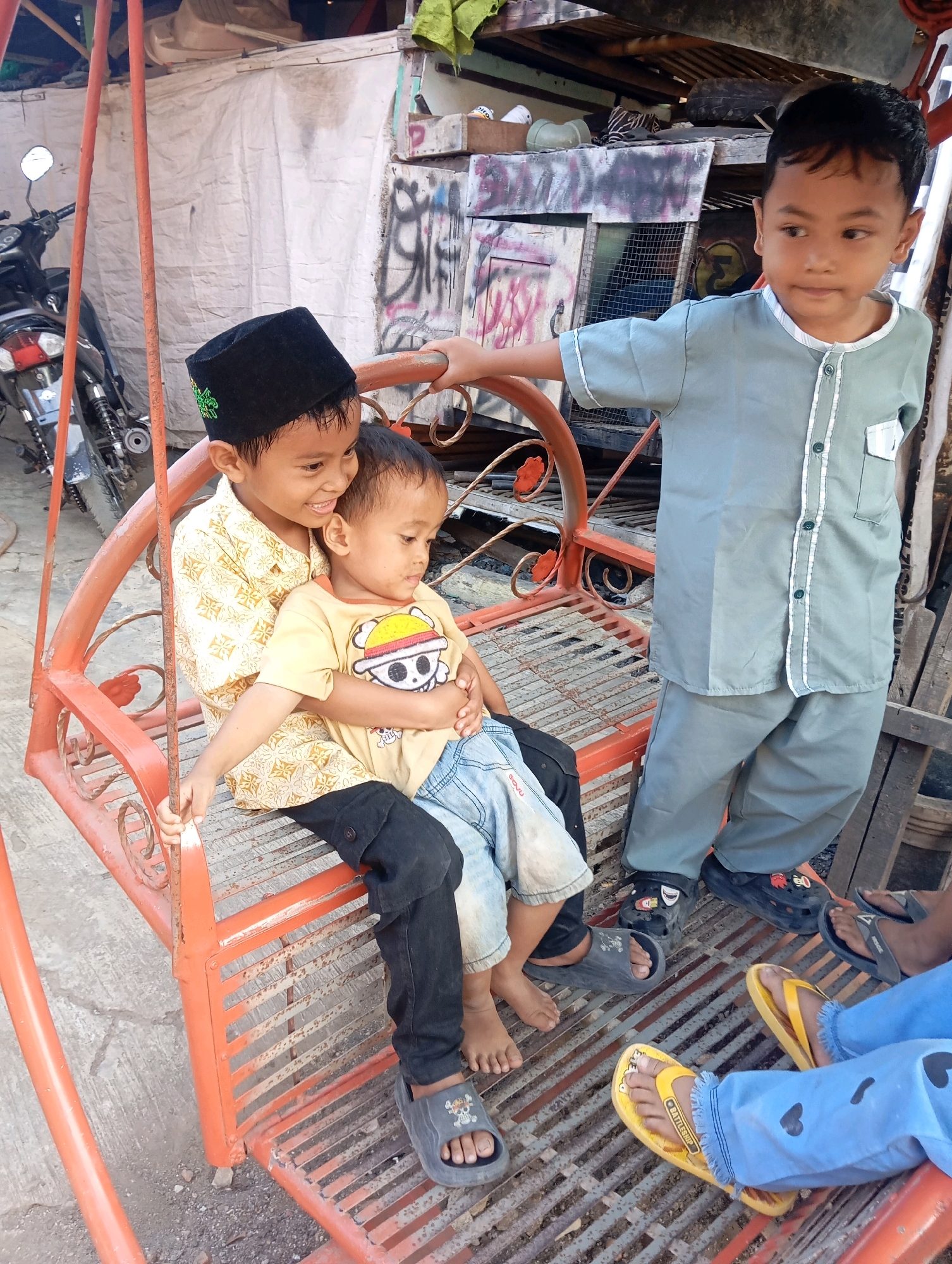 In Adi Puro, Indonesia, three young boys are depicted in a scene featuring an orange metal swing set. The boy on the far left, wearing a black peci and a patterned yellow shirt, holds the boy in the middle, who is dressed in a yellow t-shirt with a cartoon character and blue jeans, in his arms. The boy on the far right, taller than the others, stands adjacent to the swing, wearing a light blue collared shirt and matching pants. The background reveals a cluttered area with a motorcycle to the left and structures made of wood and corrugated metal, some adorned with graffiti. The ground is a mixture of concrete and gravel.