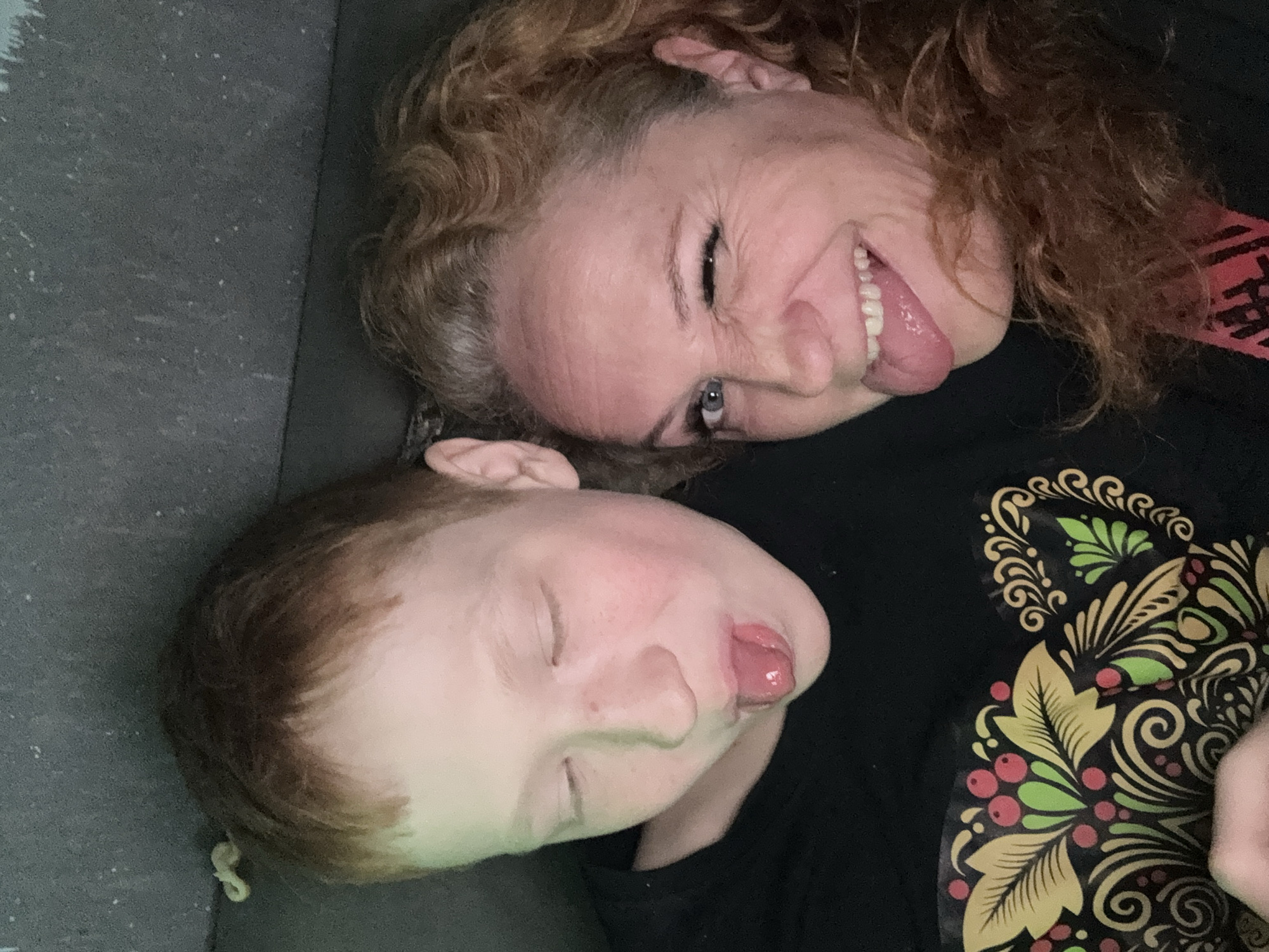 This close-up, playfully oriented portrait features an adult woman and a young child, likely taken indoors in Chaikovsky, Russia. Both subjects are lying down, with their heads close together, and are captured in the act of making funny faces by sticking out their tongues.
The adult woman, with fair skin and voluminous reddish-brown curly hair, is positioned above the child. She displays a wide smile, showing her teeth, and has pronounced wrinkles around her eyes, which appear to be squinted in amusement. Her left eye, a clear blue, is partially visible. She is wearing a dark top, with a segment of red fabric featuring partial, unreadable text visible near her hair.
Below the woman, the child has fair skin and light brown hair. Their eyes are closed or heavily squinted as they also stick out their tongue. The child's dark shirt is decorated with a vibrant, folk-art inspired pattern featuring golden swirls, green leaves, and red berries, potentially reflecting traditional Russian design elements.
The background is a simple, uniform dark gray, suggesting an indoor setting such as a couch or bed. The image conveys a strong sense of joy and lightheartedness, capturing a humorous and affectionate moment between the two individuals.