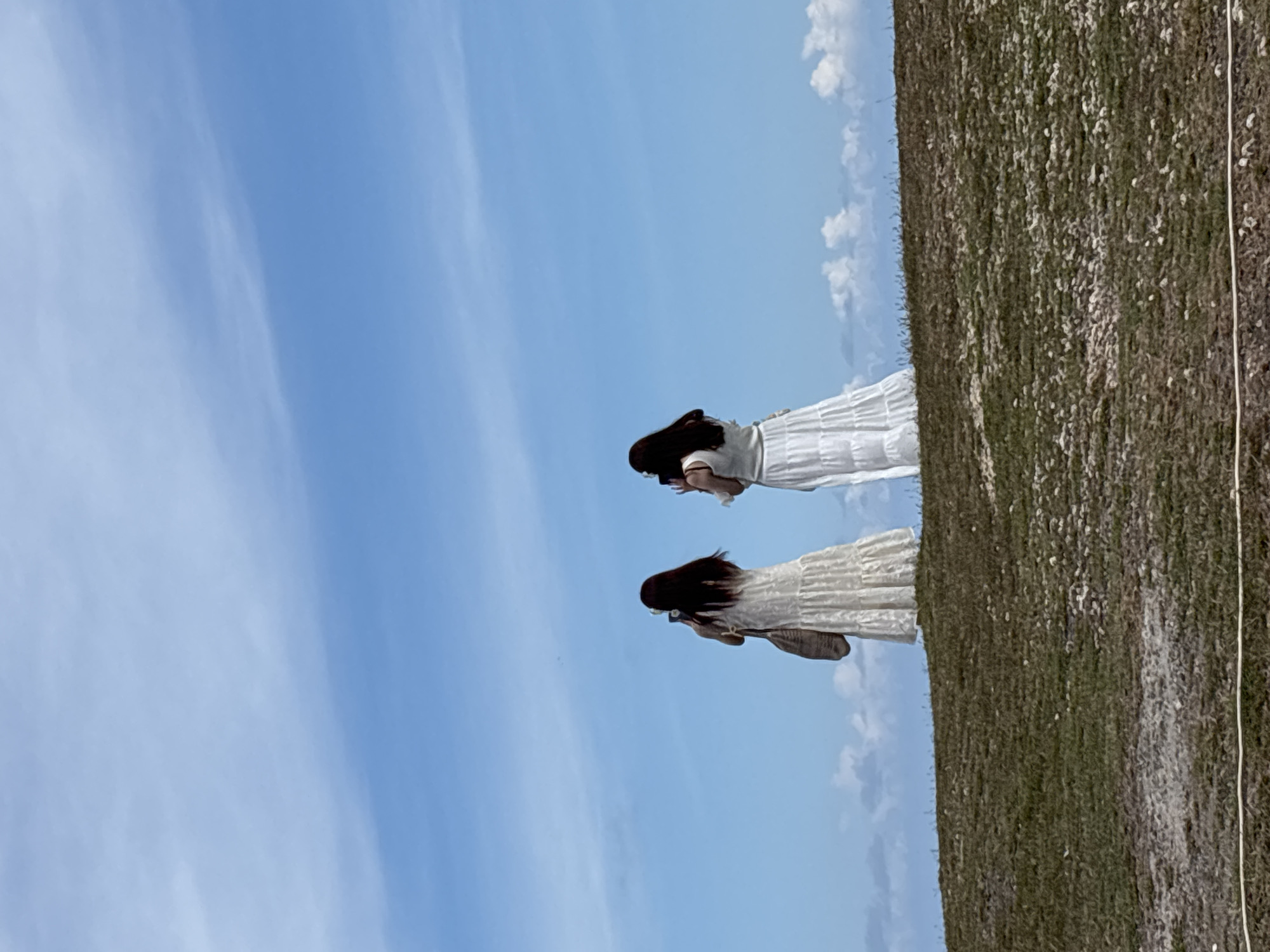 Two women with long, dark hair are standing in a grassy field under a blue sky with scattered clouds. Both are wearing white dresses. The scene is rotated counter-clockwise, placing the sky to the left and the field to the right. The specific location in Kuta Selatan, Indonesia, cannot be determined from the image alone.
