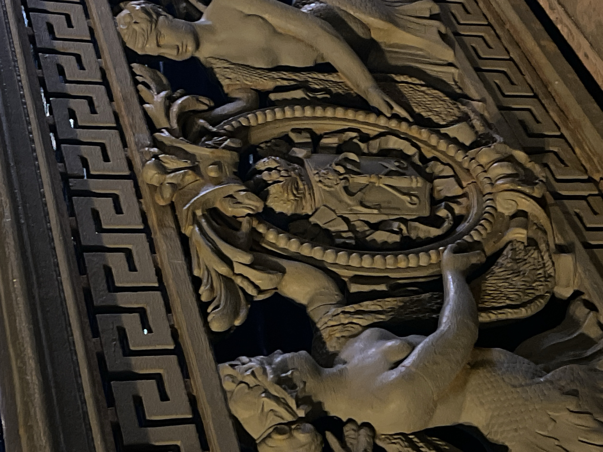 The image shows a close-up of a carved, possibly stone or plaster, architectural detail. The carving depicts two humanoid figures, likely mermaids or mermen, flanking a central oval containing what appears to be an architectural model or depiction, potentially featuring tools or instruments. The figures are arranged vertically, with one figure above the oval and the other below. A geometric pattern, reminiscent of a Greek key design, borders the left side of the carving. The scene is illuminated in such a way that highlights the texture and depth of the carving. It seems likely that this is a detail from a building in the historic center of the city of Saint Petersburg, Russia.
