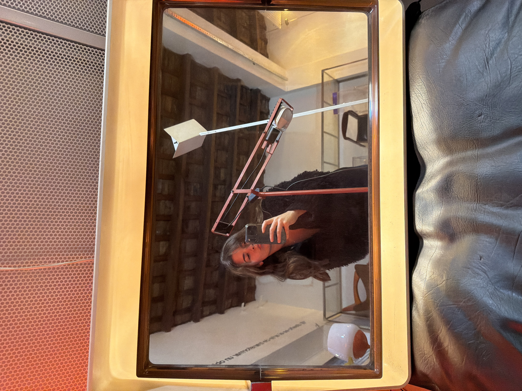 A mirror reflects a woman taking a selfie with her smartphone. The room reflected in the mirror includes a red articulated lamp, white walls with some text, and a wooden structure in the background. The mirror is surrounded by a beige frame with a brown inner border. A perforated metallic surface is visible to the left of the mirror, and a leather couch is on the right. The photograph was probably taken inside a building located in the city of Buenos Aires, Argentina, based on the text visible in the mirror.