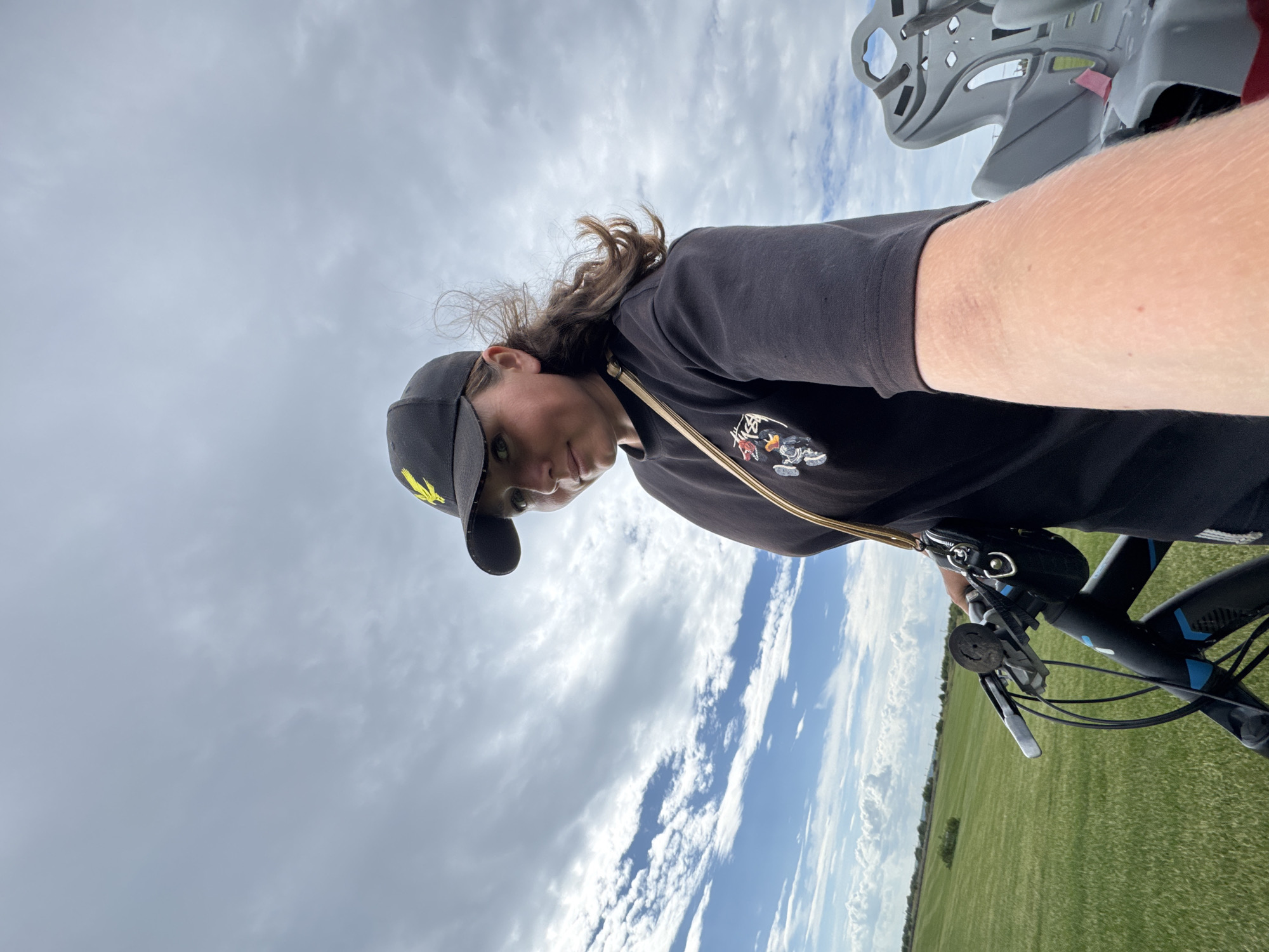 In Kempelevo, Russia, a person is standing outdoors with a bicycle. They are wearing a black baseball cap with a yellow symbol and a black t-shirt with a graphic design. A beige strap from a bag is visible across their shoulder. The background shows a cloudy sky and a vast, green field, suggesting a rural or park setting outside of the main city structures.