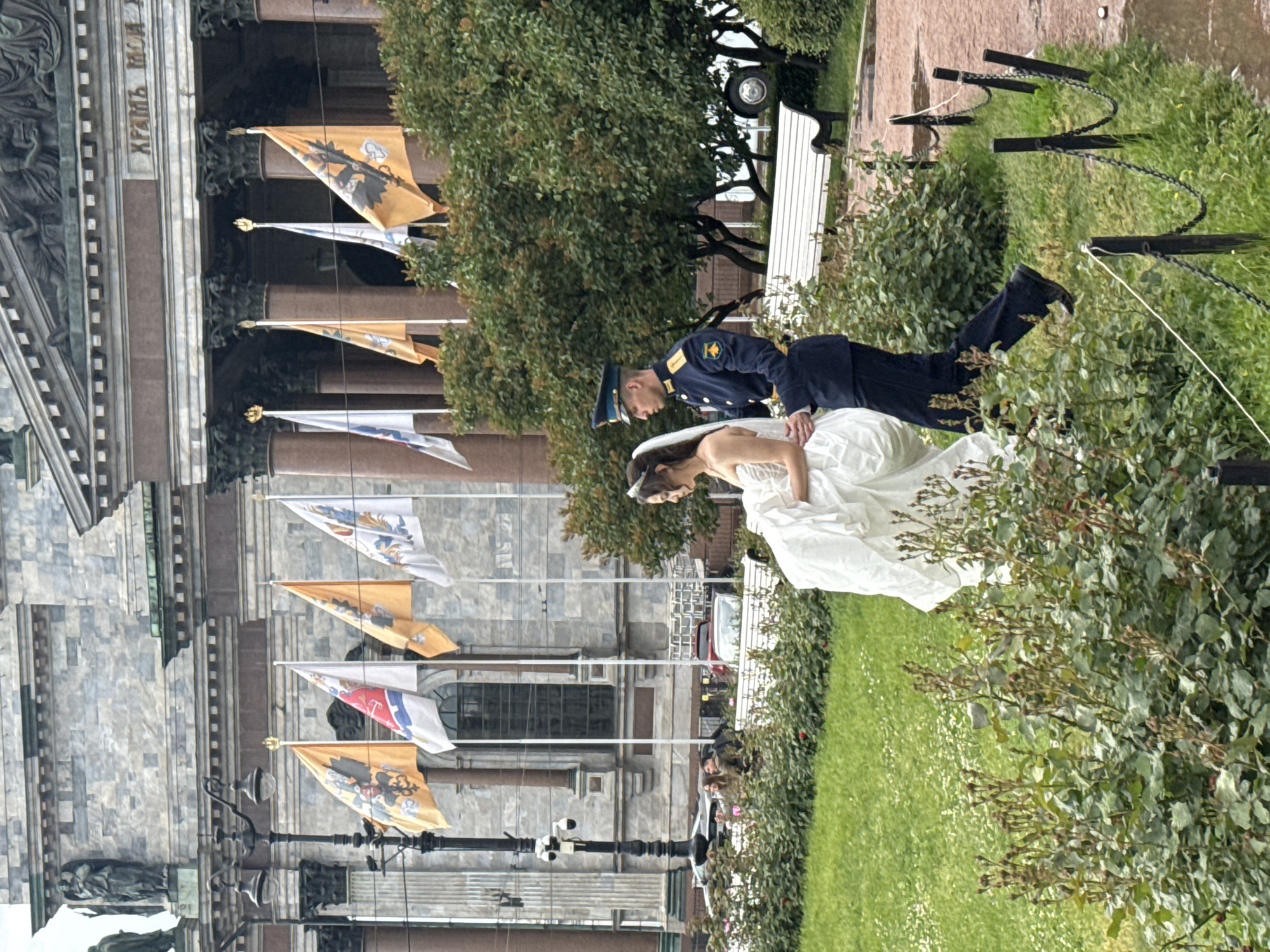 A newly married couple is walking past a large, stone building with classical columns and flags flying from poles. The bride is wearing a white wedding dress, and the groom is in a dark blue military uniform. They are in a park with green grass, trees, and rose bushes. In the background, there are cars and a white bench. The architecture of the building suggests it is a landmark in Saint Petersburg, Russia, possibly St. Isaac's Cathedral.
