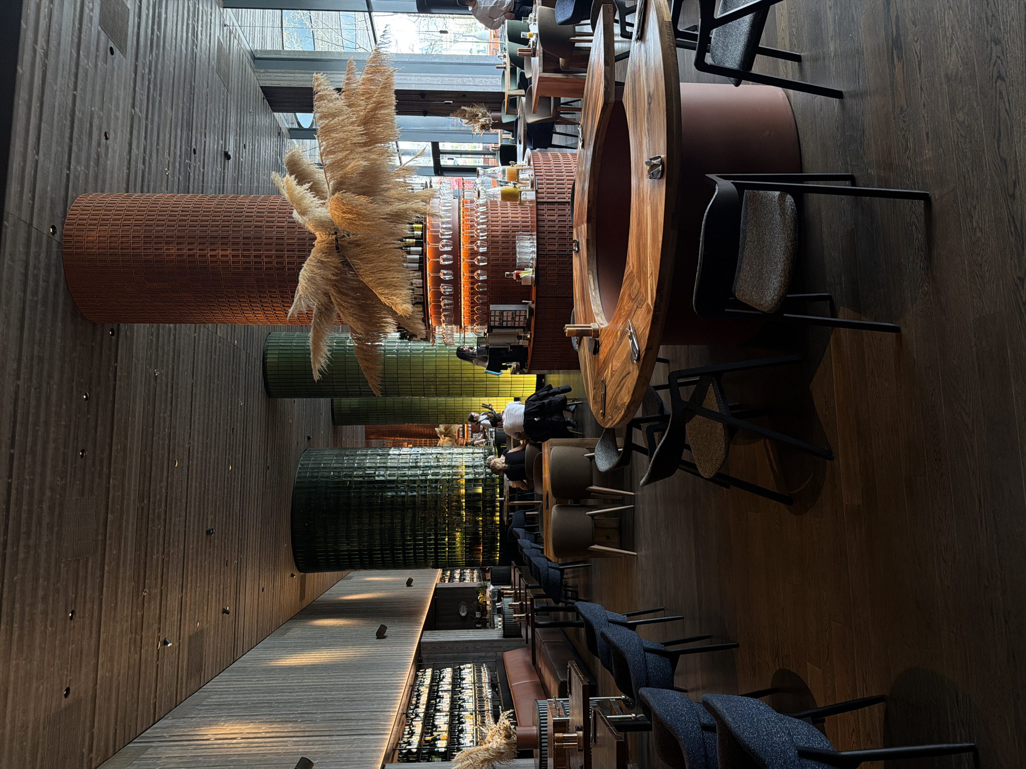 This image captures the interior of a modern and stylish restaurant or bar, located in Vynnyky, Ukraine. The space is characterized by a sophisticated design featuring rich textures, natural materials, and warm lighting.
The elongated room extends from the foreground into the background, showcasing various seating areas and a prominent bar. On the left side, large windows offer a view of outdoor trees and allow natural light to filter in. The floor is made of dark, polished wooden planks, complementing a textured, dark wooden wall that runs along the right side of the room, featuring subtle integrated lighting.
A striking focal point in the central foreground is a large, circular wooden bar counter with a polished finish. Behind it, a bar counter holds an array of drinks and glasses, suggesting active service. Integrated into the bar's structure are distinctive textured cylindrical columns. The most prominent one, in the foreground, is a burnt orange or terracotta color with a mesh-like pattern, dramatically adorned with large, fluffy, light-colored plumes, likely pampas grass. Another similar cylindrical structure, further back, is visible in a dark green hue.
Seating arrangements vary throughout the room. In the foreground, rows of chairs with dark frames and speckled blue upholstery are arranged, indicating a lounge or waiting area. Further into the room, there are tables paired with modern chairs, some featuring light grey upholstered seats, and sections of brown leather-like booth seating. In the distant background, a large, illuminated display cabinet filled with numerous bottles, likely wine or spirits, is visible.
Several people are present in the scene, contributing to a lively but relaxed atmosphere. A person, presumably staff, is seen behind the bar. Other patrons are scattered across the seating areas, either seated at tables or standing near the bar, engaging in casual dining or socializing. The combination of natural light and warm artificial illumination suggests the time of day is likely late afternoon or early evening. No discernible text is visible in the image.