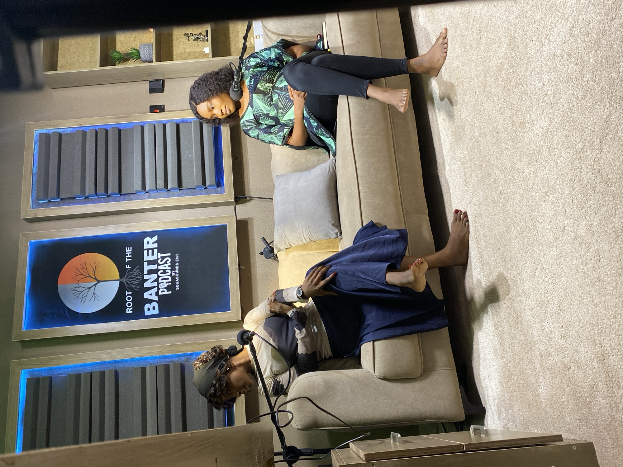 The image shows two women in a podcast studio, likely in Gura Top, Nigeria. Both subjects are seated on a tan couch, facing towards each other. The woman on the left is wearing a green and black patterned top and black leggings, with headphones on. She is looking down at her phone. The woman on the right is wearing a long navy blue skirt and a patterned top. She is also wearing headphones and appears to be talking into a microphone, suggesting they are recording a podcast.
The studio has soundproofing panels on the walls, with blue accent lighting. A poster for "ROOT OF THE BANTER PODCAST BY SUKASOUNDS ENT" is visible on the wall. The floor is carpeted in a light beige color. The lighting suggests it is daytime, as shadows are cast on the floor. The overall atmosphere seems relaxed and professional, conducive to recording a podcast.