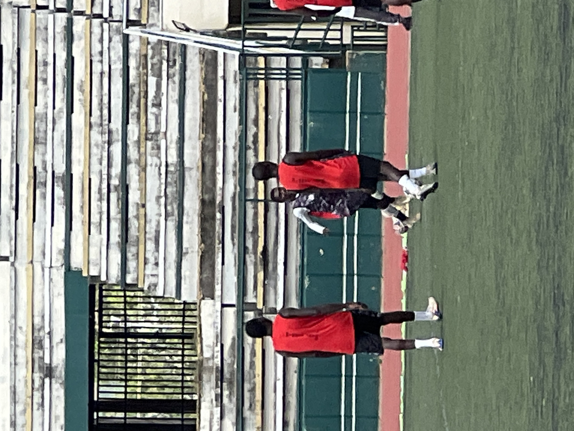 The image shows a group of men on a green turf field, likely a sports field, at the New Stadium in Nigeria. In the foreground, a man in a red sleeveless shirt and black shorts stands facing away from the camera. To his left, another man in a camouflage-patterned shirt and black shorts is running. In the mid-ground, a third man in a red sleeveless shirt and black shorts is also facing away from the camera, and further to the right, another person in a red shirt is partially visible. The background features tiered stadium seating constructed with what appears to be weathered materials. The field has a red track running along its perimeter.