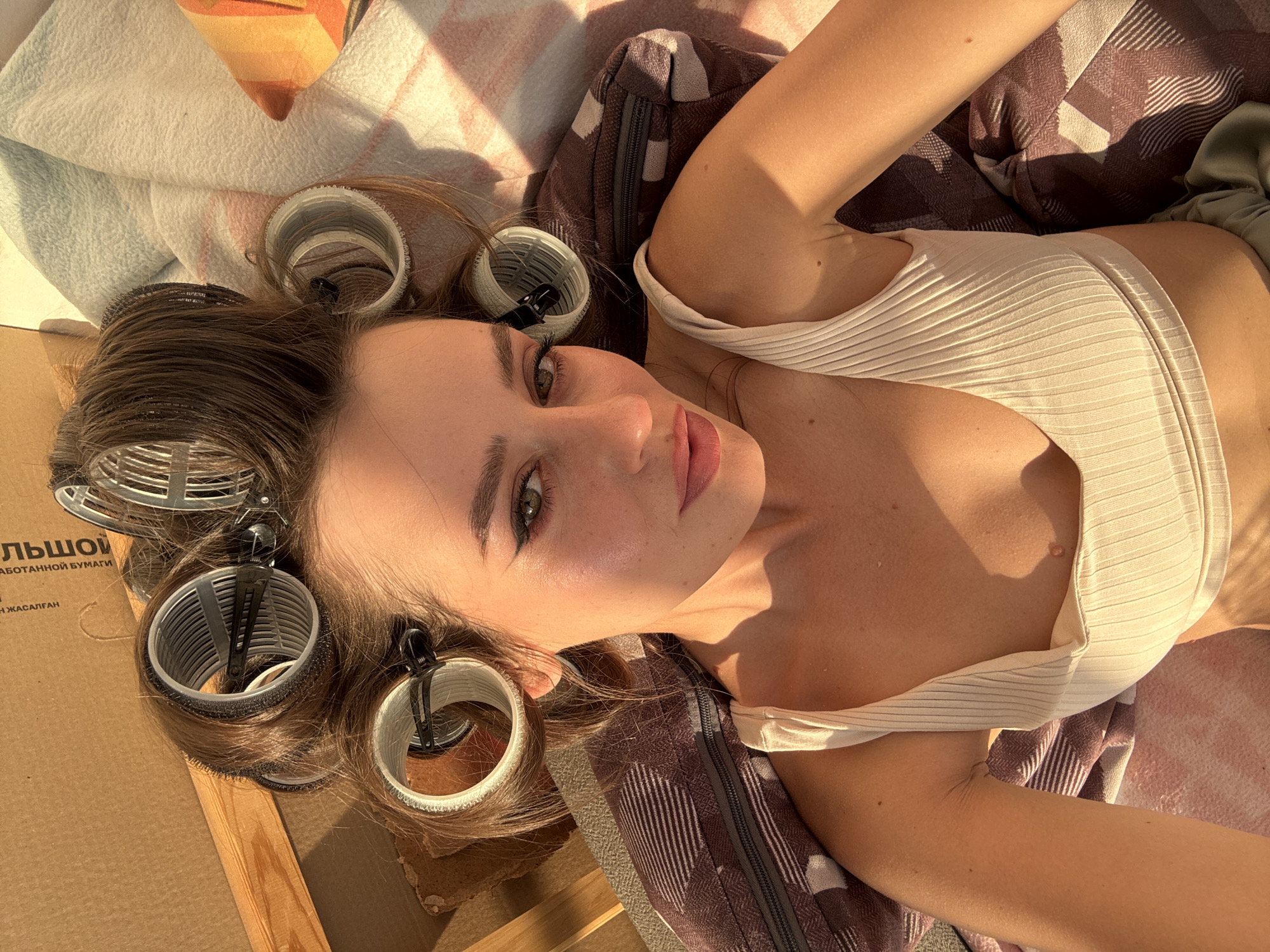 A top-down, slightly tilted image captures a young woman lying on her back, looking directly at the camera. Her hair is styled with several large, grey hair rollers secured with black clips, visible around her head. She has dark brown hair, green eyes with winged eyeliner, and light pink lips. Her skin is fair, with some freckles visible on her nose and cheeks.
She is wearing a light beige or cream-colored ribbed crop top that reveals her midriff. Her right arm is raised and bent, with her hand out of frame, and her left arm is positioned alongside her body. Sunlight casts strong shadows, suggesting a bright sunny day. The lighting illuminates her face and upper body, creating warm tones.
She is lying on what appears to be a patterned blanket or cushion in shades of brown and white, and possibly another light-colored blanket to her left. To her left, part of a cardboard box is visible, with Cyrillic text that appears to read "ЛЬШОЙ" (likely part of "БОЛЬШОЙ" meaning "big" or "large") and "АБОТАННОЙ БУМАГИ" (meaning "processed paper") along with "ЖАСАЛҒАН" (Kazakh for "made"). This suggests the presence of packaging or a box nearby. The overall setting appears to be casual and indoors or in a sheltered outdoor space, given the presence of the cardboard box. Her expression is serene and she appears to be relaxed.