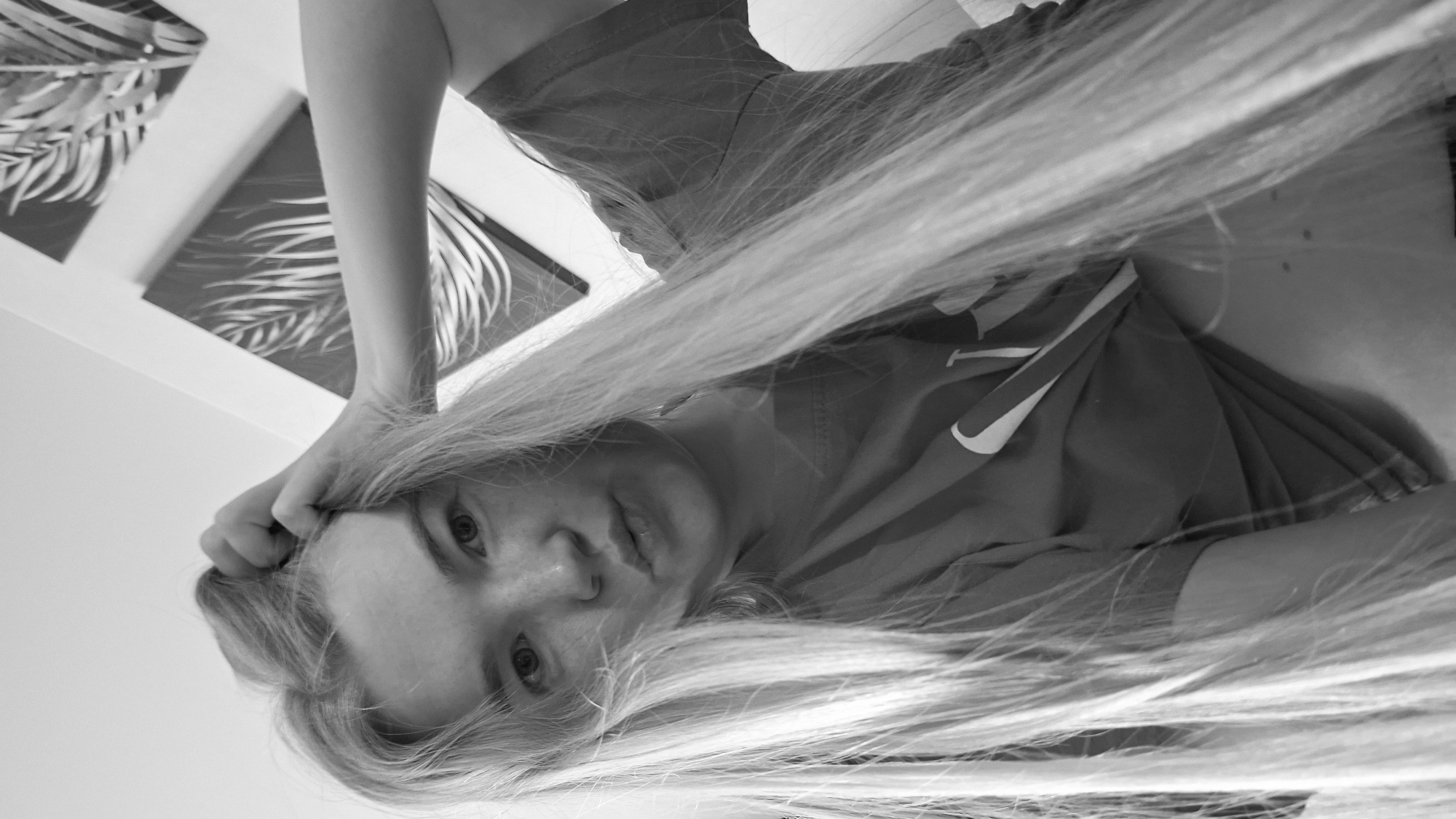A black and white close-up photograph depicts a young, fair-skinned person with light-colored, long hair partially obscuring the view. The person is wearing a t-shirt with a visible swoosh logo. The person's left hand is near their face, possibly touching their hair. Three framed pictures displaying leaf patterns are on the wall behind the person. The location is presumed to be in Chaikovsky, Russia.