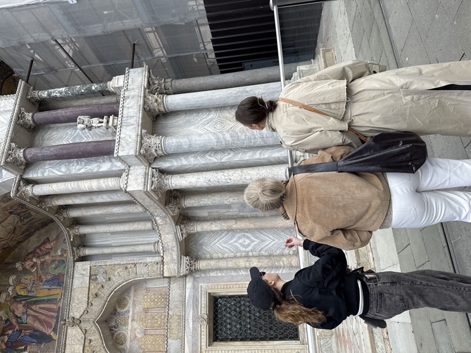 The image depicts three individuals standing on a paved surface, looking upwards at the ornate facade of St. Mark's Basilica in Venice, Italy.
From left to right:
1. A female individual, viewed from the rear-right, wears a black baseball cap, black sunglasses, a black bomber jacket, dark grey wide-leg jeans, and a white fanny pack. Her light brown hair is pulled into a ponytail beneath the cap. Her right arm is slightly raised with fingers splayed, gesturing towards the building.
2. A female individual, viewed from the rear, wears a tan suede-like short jacket, white trousers, and a patterned scarf visible around her neck. Her blonde hair is styled in a bun. A large, dark brown or black shoulder bag hangs from her left shoulder.
3. A female individual, viewed from the rear-left, wears a light beige long trench coat. Her dark hair is pulled back into a low bun or ponytail.
All three individuals are focused on the elaborate marble and mosaic facade of the basilica, which dominates the background. The building features multiple tiers of classical columns with intricate Corinthian capitals, constructed from various types of marble, including light grey/white veined marble and darker reddish-purple columns. A segment of a multi-colored mosaic depicting multiple figures is visible above an archway on the upper left. Below this, an arched window with a dark grate is present. On an upper tier to the right, a small white statue of a figure carrying an object stands on a ledge. To the far right, a section of the building is obscured by scaffolding draped in white protective sheeting, indicating ongoing work. A dark barrier or fence is visible at the bottom right. The ground beneath the individuals is a light-colored paved stone surface.
