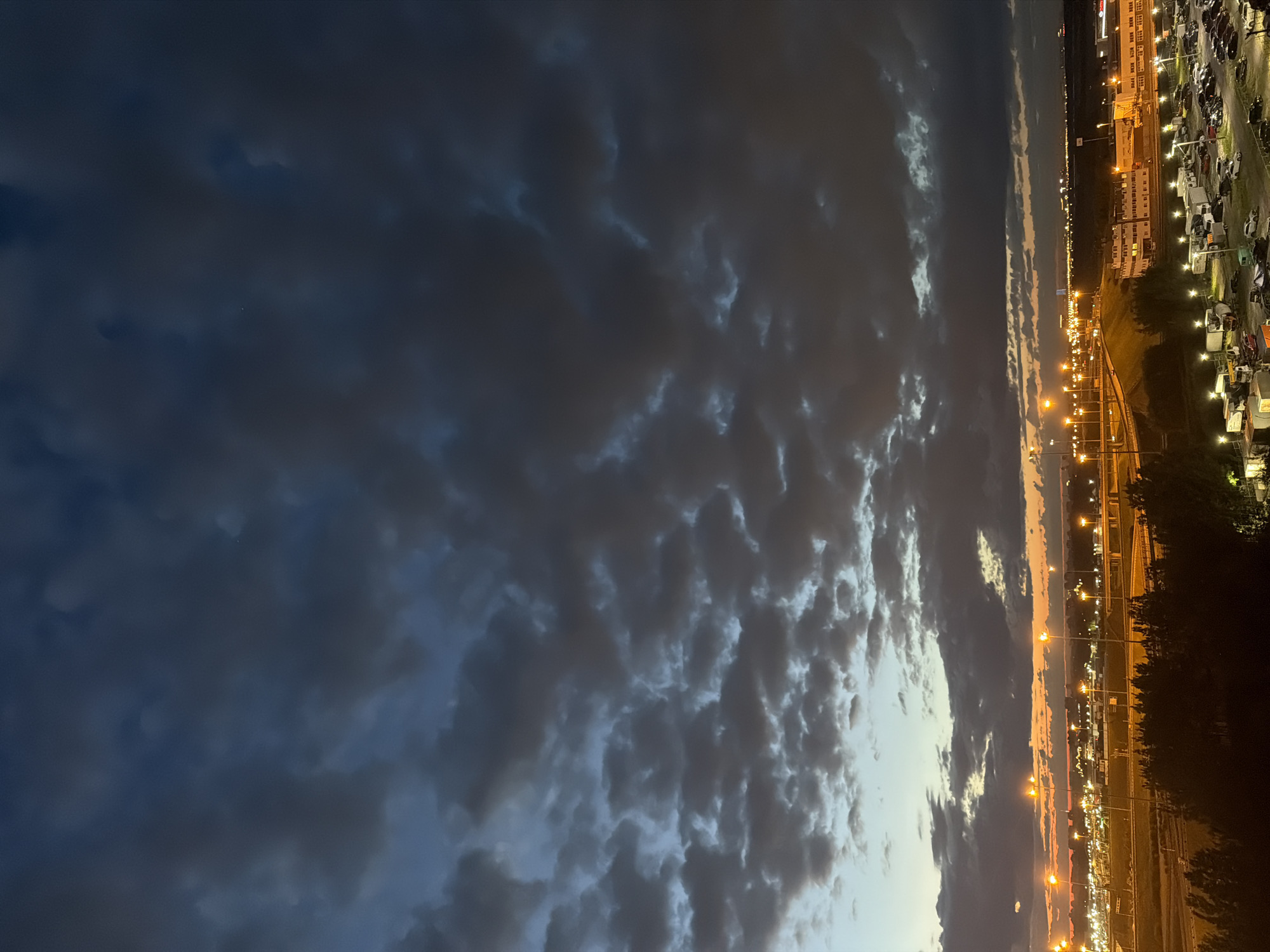 The image captures a twilight scene over a city. The sky is dominated by dark, dramatic clouds, with patches of lighter blue and orange peeking through near the horizon, suggesting the last remnants of sunset. Below the sky, a sprawling urban landscape is illuminated by numerous streetlights and building lights. The cityscape features a complex network of roads and bridges, as well as various industrial or commercial buildings. In the foreground, a dense line of dark trees obscures the ground level, and a parking lot filled with cars and trucks is visible, indicating a busy area. There are no people clearly visible in the frame.