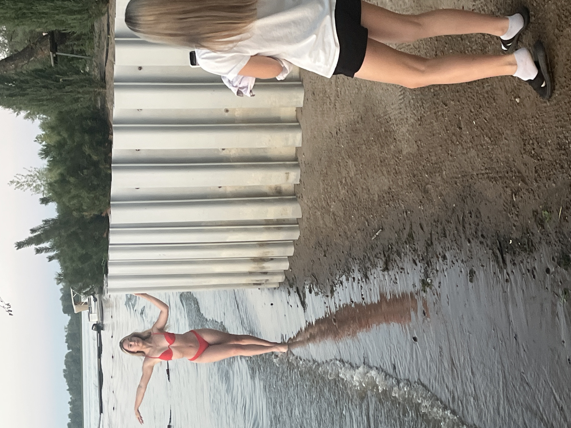 In Shumeika, Russia, on a beach by a body of water, a young woman in a red bikini stands in shallow water with her arms extended, facing away from the camera. Her reflection is visible on the wet sand. Behind her, a ribbed metal barrier runs horizontally across the frame. In the foreground, another young woman, seen from the back, holds a phone and a towel, wearing a white t-shirt, black shorts, and socks with sandals. The background shows trees and a small boat on the water. The sky is overcast.