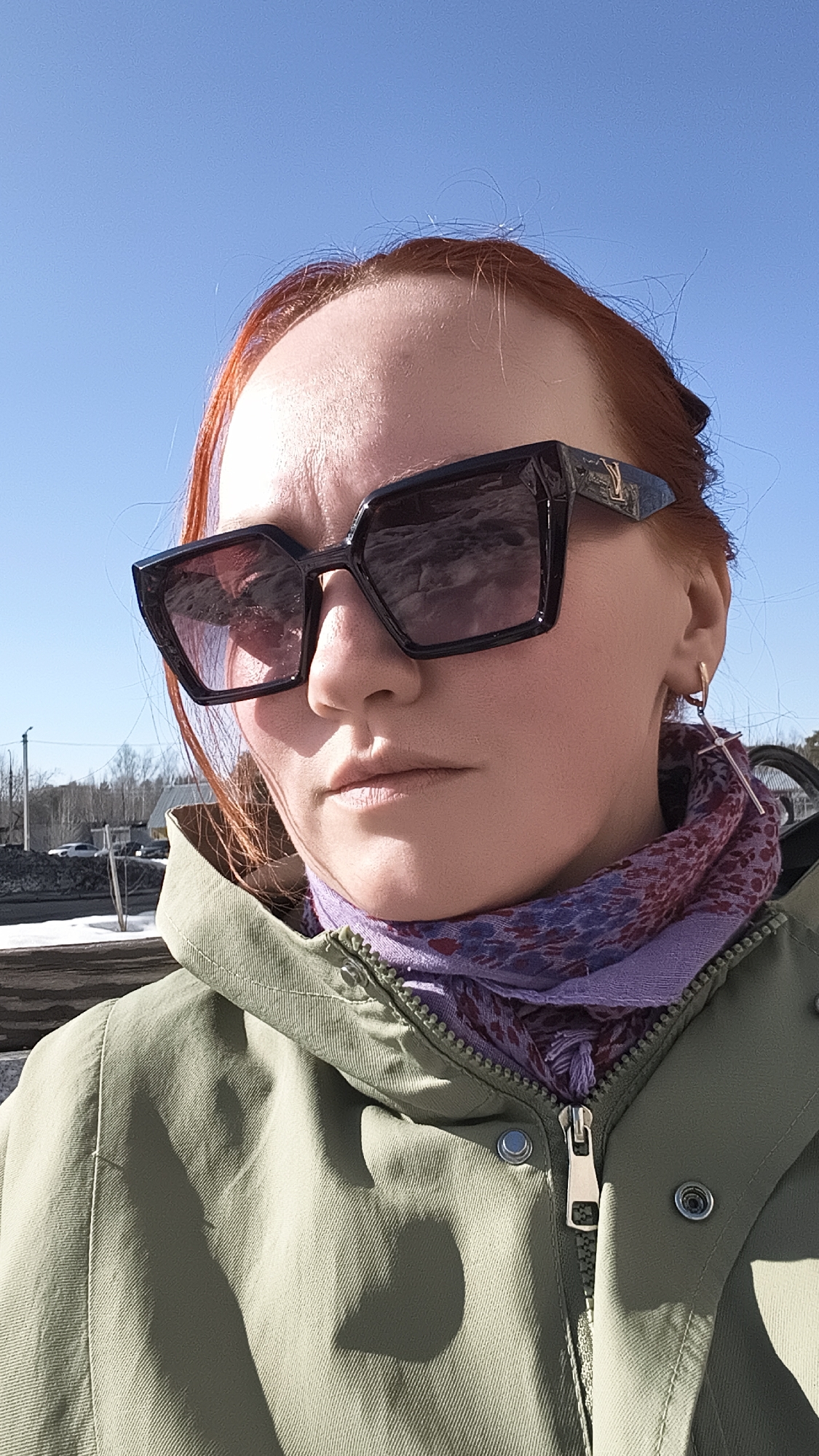 A selfie of a fair-skinned woman with auburn hair and large black sunglasses, wearing a green jacket and patterned purple scarf. She has a cross earring in her left ear. In the background is a fence and some trees under a clear blue sky. The location is indicated as Chaikovsky, Russia, but there is no specific information in the image to confirm that.