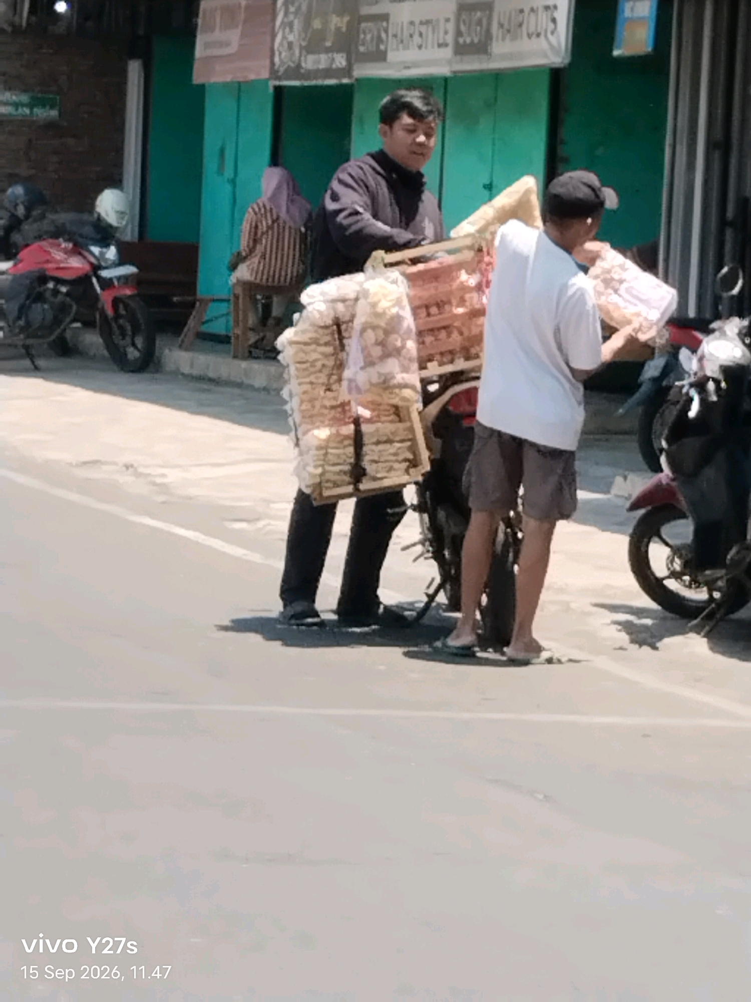 The image depicts a street scene in Sukorejo, Indonesia. Two men are visible, one on the left wearing a dark jacket and the other on the right wearing a white t-shirt and shorts. They appear to be associated with a motorcycle that is loaded with a large quantity of packaged goods, likely snacks or food items, strapped to its rear. The man on the right is holding a package. In the background, there is a building with green doors, and a red motorcycle is parked to the left. A person wearing a headscarf is seated on a stool near the building. The overall environment suggests a commercial or roadside setting with pedestrian and vehicular traffic. The lighting indicates it is daytime.