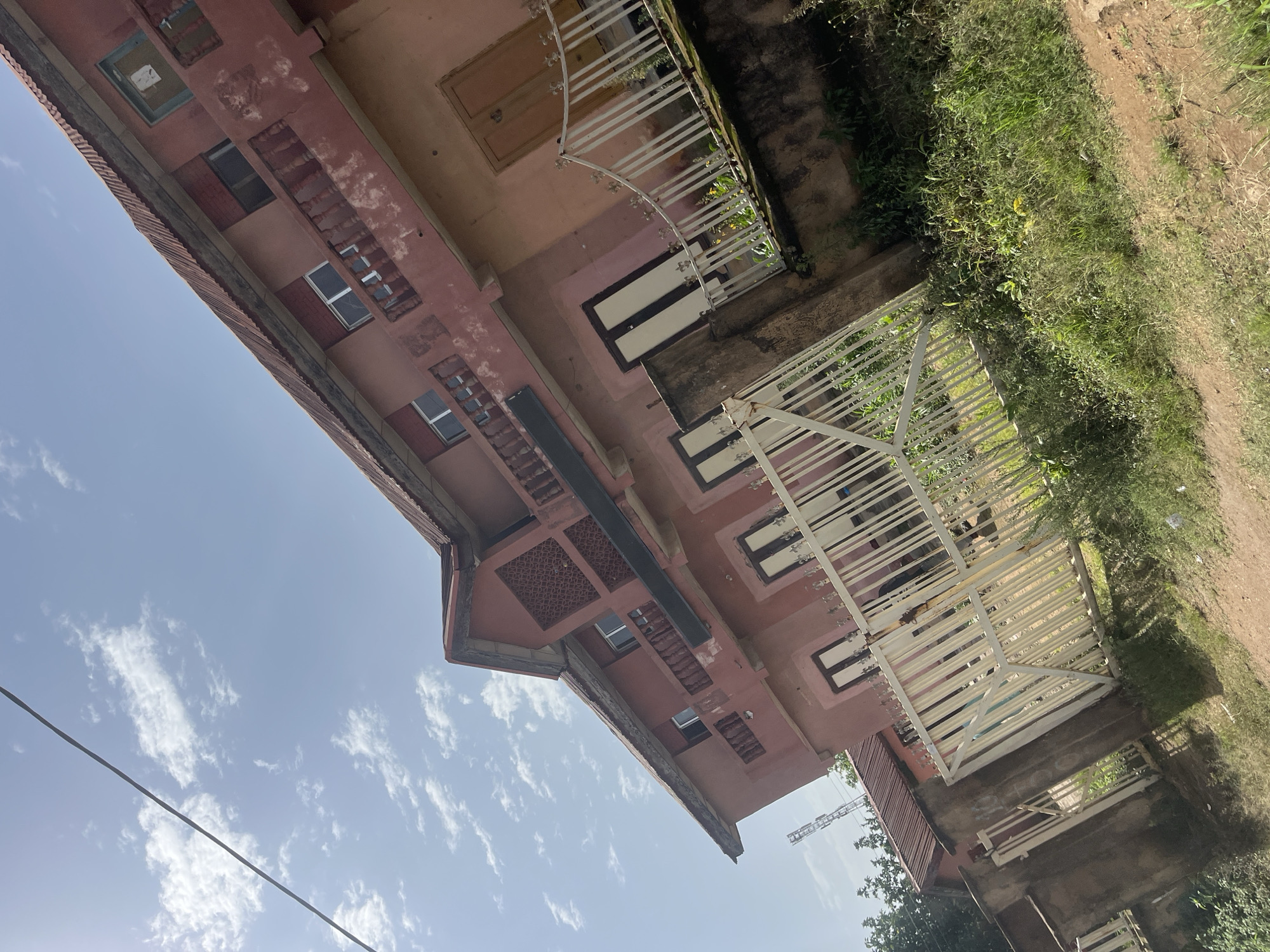 The image depicts a multi-story, salmon-pink building with a rusted corrugated metal roof, located in Bukuru, Nigeria. The building appears somewhat aged, with peeling paint and weathered balconies. There are several windows and doors visible, some with wooden frames and others with glass panes. A white metal gate and fence front the property, with overgrown green vegetation and grass surrounding it. The sky above is a light blue with scattered white clouds, and a power line runs diagonally across the upper portion of the frame. A cell tower is visible in the distance to the left of the building. The perspective is a low-angle shot looking up at the building.