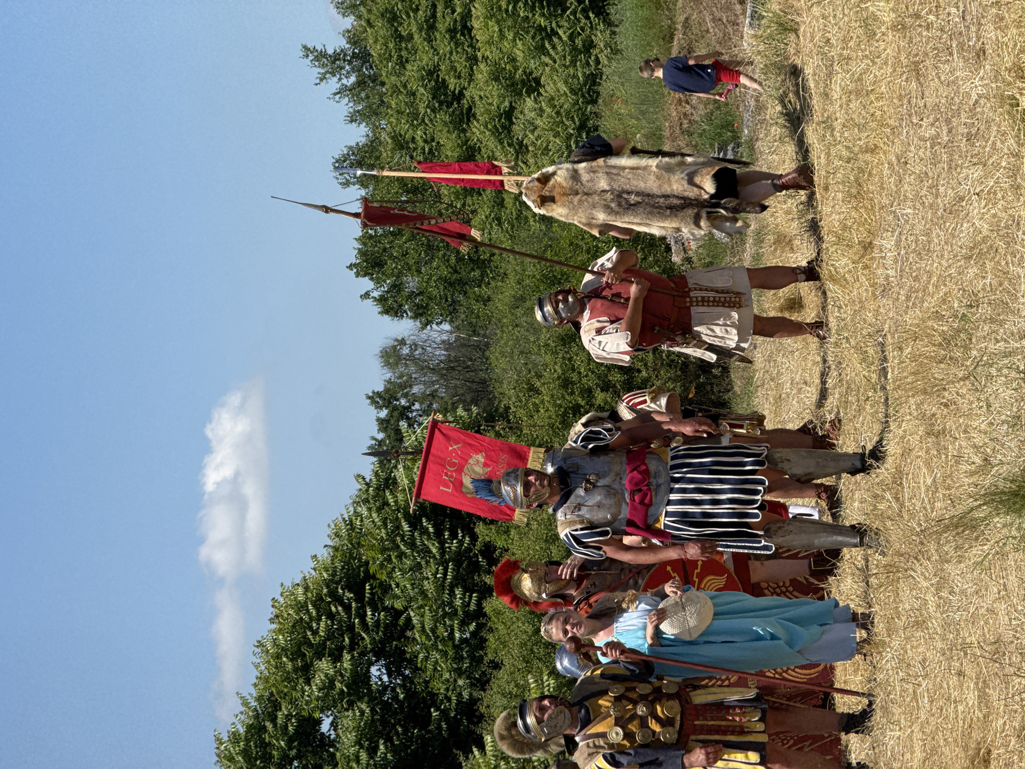 The image depicts a historical reenactment occurring outdoors in a dry, grassy field, presumably in Gigen, Bulgaria, under a clear blue sky with sparse white clouds.
Seven individuals are prominently featured, primarily dressed in Roman-era military and civilian attire. From left to right:
1. A male figure in elaborate Roman legionary armor, featuring a gold and dark blue segmented chest piece, a golden-yellow and dark blue striped skirt, greaves, and a helmet topped with a brown fur crest. He holds a long, brown wooden staff vertically.
2. A female figure in a light blue toga-like dress, holding a white, circular woven object (possibly a fan or small basket) in her right hand. She looks towards the right.
3. A male figure partially obscured behind the woman, wearing Roman armor with a distinctive red-plumed helmet and a red tunic. A large, round red shield with a central gold emblem is visible at his side.
4. A central male figure, likely a Roman standard-bearer, wears a silver-colored breastplate with blue shoulder pieces, a white and blue vertically striped skirt, and greaves. His helmet features a blue feather crest. He firmly holds a red rectangular vexillum (banner) displaying "LEG X" in white letters above a white boar emblem, affixed to a tall pole.
5. A male figure in a red tunic and silver breastplate, with a white-painted face and a helmet, holds a tall spear or standard-like pole.
6. A male figure, seen from the back, wears a large brown fur cape over a dark garment, typical of a standard-bearer or officer. He holds a pole with a smaller red flag or banner attached, which is partially obscured.
7. In the far right background, a young child, a boy in a dark blue t-shirt and red shorts, walks across the grassy field away from the group, holding a small red object.
The ground is covered with sparse, dry, light brown grass and some greener patches. The background consists of dense green deciduous trees under the bright midday sun. No active interaction or combat is depicted; the costumed individuals appear to be standing in formation or posing.