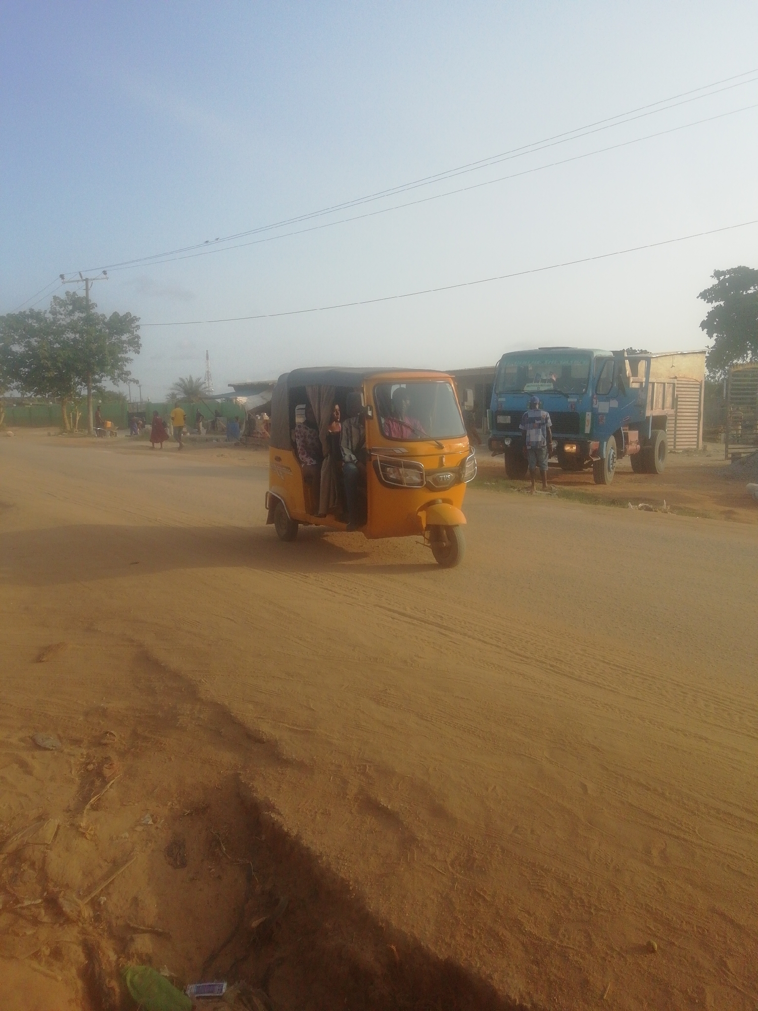 The image depicts a street scene in an unidentified location in Unknown, Unknown. The environment appears dusty and sunlit. A yellow auto-rickshaw, with several visible passengers inside, occupies the center foreground. To the right of the auto-rickshaw, a blue truck is parked, with a man standing near its front. In the background, several people are walking and standing near buildings. Overhead, power lines extend across the sky. A tree is visible in the distance. The road surface is unpaved and sandy, with tire tracks visible.