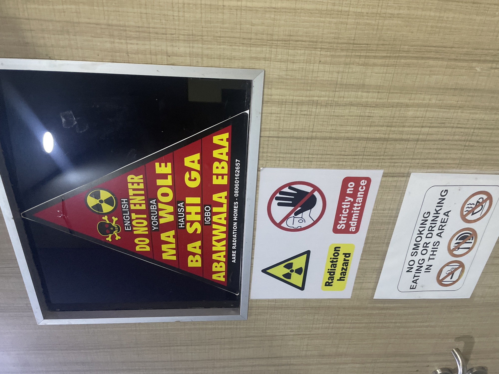 The image shows a closed door with several signs posted on it. At the top, a large triangular sign with a red background and black border warns of radiation hazard with a skull and crossbones, a radiation symbol, and text in English, Yoruba, Hausa, and Igbo languages stating "DO NOT ENTER" and its translations. Below this, two smaller rectangular signs are visible. The left sign has a yellow background with a black radiation symbol and the text "Radiation hazard". The right sign has a white background with a red border and depicts a hand in a circle with a diagonal line through it, along with the text "Strictly no admittance". At the bottom, a rectangular sign with a white background displays "NO SMOKING EATING OR DRINKING IN THIS AREA" along with three prohibition symbols: no smoking, no eating, and no drinking. The door appears to be made of wood with a light wood grain pattern, and a silver door handle is visible on the left side. The environment suggests an indoor setting, likely a facility or building where radiation is present. The signs indicate a restricted area due to radiation, with prohibitions on entry, smoking, eating, and drinking. The presence of multiple languages on the main sign suggests the location is in a diverse linguistic region. Given the information on the top sign, "Aare Radiation Homes", and the context of the signs, this door is located in Abuja, Nigeria, specifically within an establishment related to radiation services or housing.