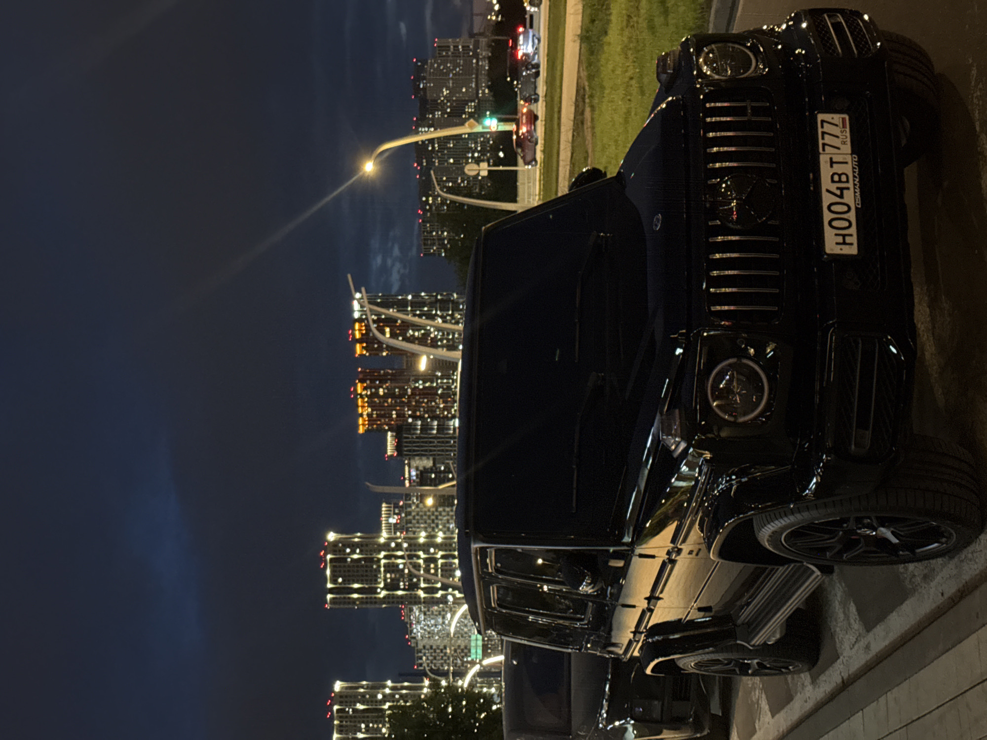 The image depicts a black Mercedes-Benz G-Class parked on a street in Khimki, Russia, at night. The license plate number is H004BT 777. In the background, illuminated high-rise buildings are visible against a dark sky. A street lamp casts light on the area near the car. There are other vehicles on the road.