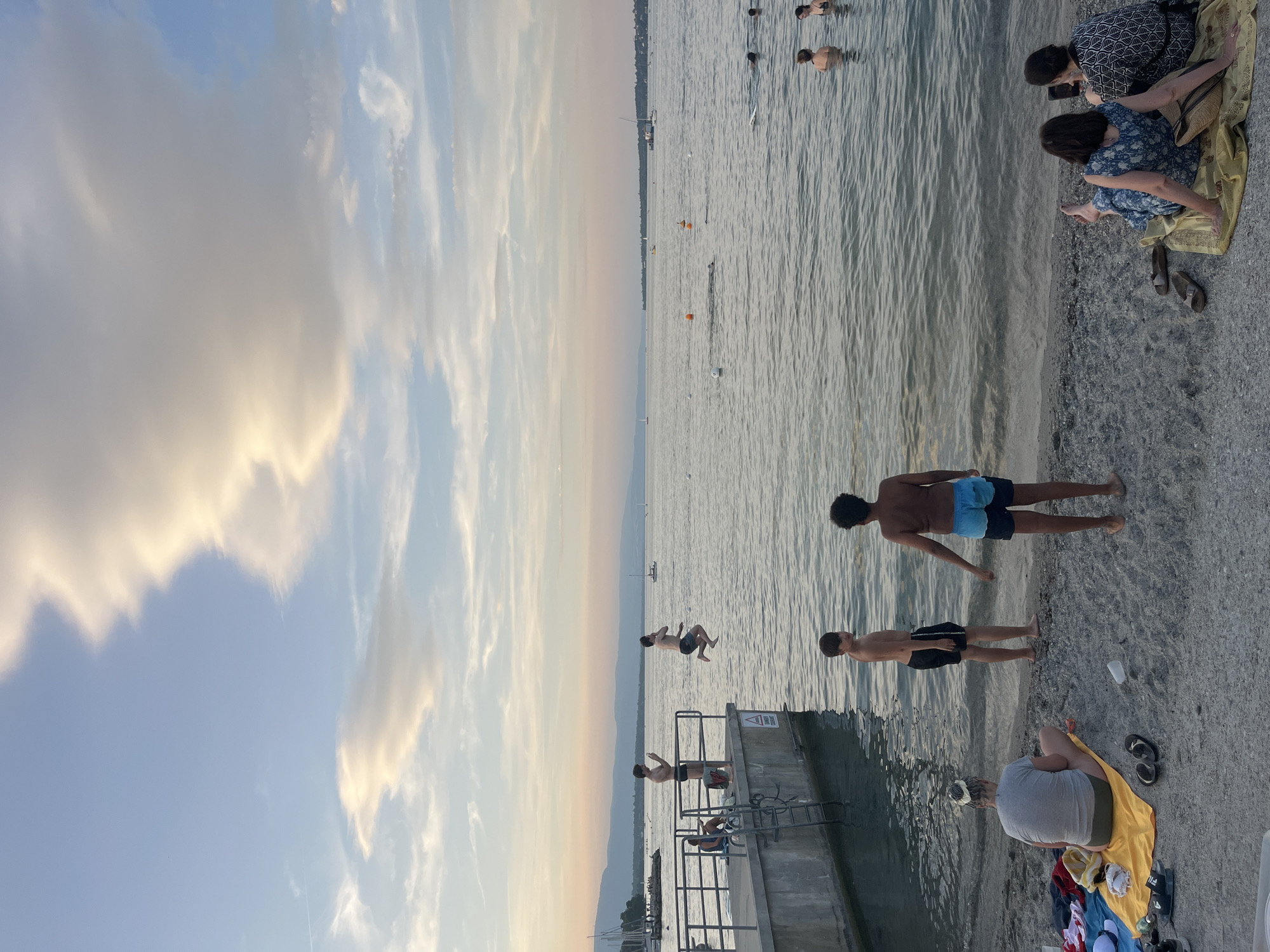 This image captures a vibrant summer scene at Plage des Eaux-Vives in Geneva, Switzerland. The golden hour light casts a warm glow over the placid waters of Lake Geneva, with the distant Jura mountains forming a serene backdrop. In the foreground, a sandy beach is dotted with people enjoying the water and the sunset.
On the beach, a woman in a grey t-shirt and green shorts sits on a yellow towel, facing the lake. Next to her, two young boys, one in black swim trunks and the other in blue swim trunks, stand near the water's edge, facing away from the camera. Further down the beach, two women are seated on a yellow blanket, one in a blue floral dress and the other in a black and white patterned top.
A concrete pier extends into the lake, where several individuals are visible. One person is captured mid-air, leaping into the water from the pier. Another person stands on the pier, seemingly taking a photo with their phone. A younger person sits on a foldable chair near the edge of the pier.
The lake itself is populated with swimmers and several buoys marking swimming areas. Sailboats and other vessels dot the horizon, adding to the idyllic atmosphere. The sky above is a canvas of soft blue, with wispy clouds tinged with the warm hues of the setting sun.