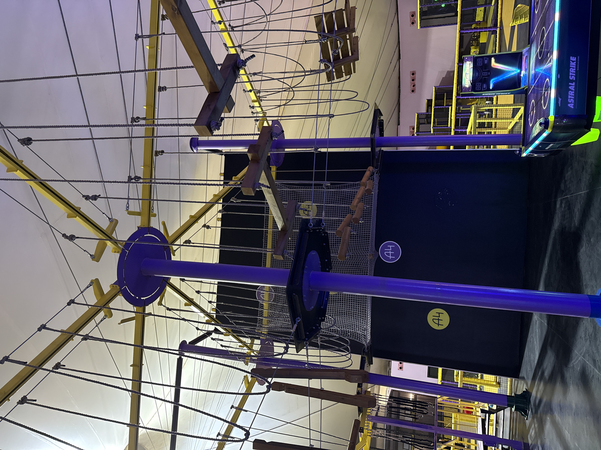 The image shows an indoor ropes course and entertainment center, likely a family fun park or similar venue. In the foreground, an "Astral Strike" air hockey table is visible, lit with neon blue and green lights. Behind the air hockey table, a tall, multi-level ropes course structure dominates the scene. The ropes course features wooden obstacles, netting, and purple and yellow support beams and poles. The walls of the venue are dark blue with some visible circular markers labeled "A4". In the background, more sections of the ropes course and other play structures are visible, also with yellow accents. The lighting appears to be a mix of artificial light, creating a vibrant and colorful atmosphere. The city of Bodrum, Turkey is not depicted or referenced in the image. There are no people visible in the image.