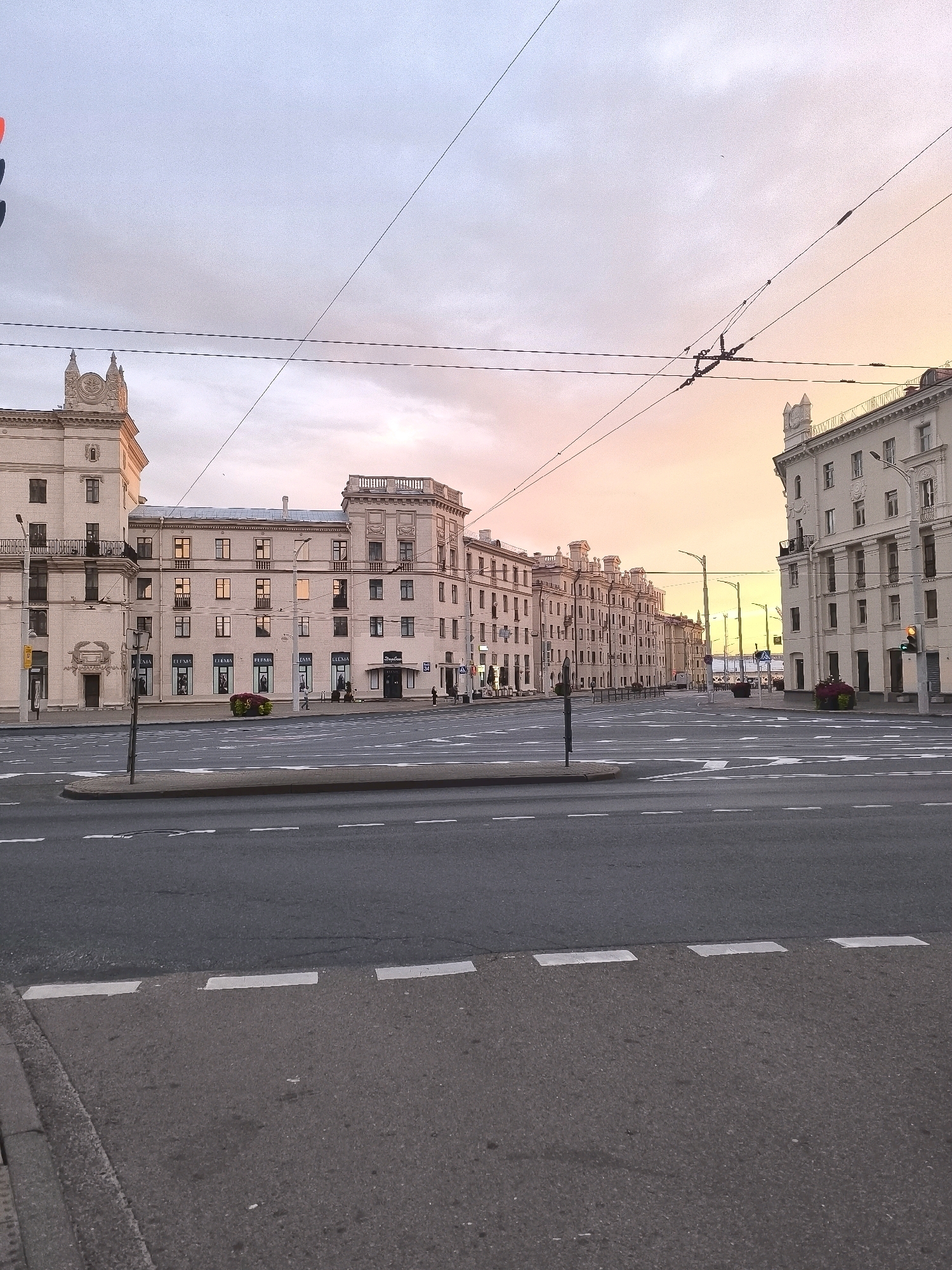 The image captures a wide-angle view of an empty intersection in Minsk, Belarus, during what appears to be sunset or sunrise. The sky is painted with soft hues of pink, orange, and blue. Large, light-colored buildings with neoclassical architectural elements line both sides of the street. These buildings have multiple stories, many windows, and ornate decorative details.
In the foreground, a paved road with clear lane markings and directional arrows is visible, indicating a multi-lane intersection. There are no vehicles or pedestrians present, creating a sense of stillness and emptiness. Overhead, a complex network of electrical wires, likely for public transport like trams or trolleybuses, stretches across the sky. A traffic light, showing a red signal, is visible on the left side of the frame. Flower arrangements in planters are placed near the building entrances. The overall impression is of a grand, but deserted, urban setting.
