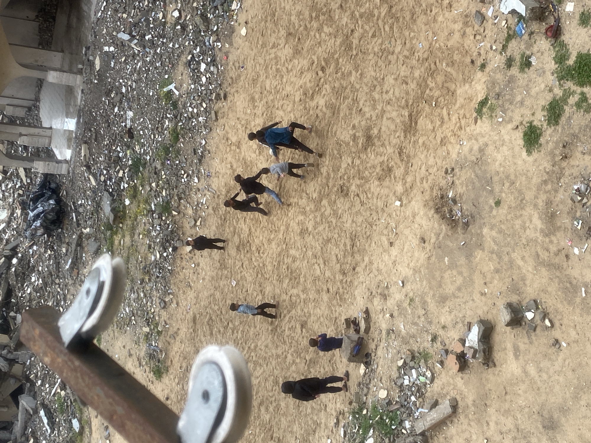 An elevated view captures a group of approximately nine male individuals, appearing to be children or young adults, playing on a sandy, debris-strewn ground in Khan Yunis, Palestinian Territory. In the central-right area, seven individuals are actively engaged in a game, likely soccer, with several figures in motion. One individual near the center wears a blue jacket; others are in dark clothing. To the left, one individual stands observing, while another is seated on a concrete block beside a small cluster of sandals. In the upper-middle, a solitary figure stands apart. The ground surface is predominantly light brown sand/dirt, interspersed with small patches of green vegetation and scattered debris, including concrete fragments and miscellaneous refuse.
The background reveals an extensive landscape of urban destruction, characterized by large piles of rubble, fragmented concrete, and exposed rebar, suggesting demolished or severely damaged structures. Towards the upper right, a section of a building with multiple arched openings and support columns remains partially standing, with puddles visible on its interior floor. A dark, possibly tarp-covered mound is situated among the rubble in the upper-left background. In the immediate foreground, a rusty, rectangular metal beam with two white pulleys attached descends from the top-left, partially framing the scene, indicating the photograph was taken from an elevated position.